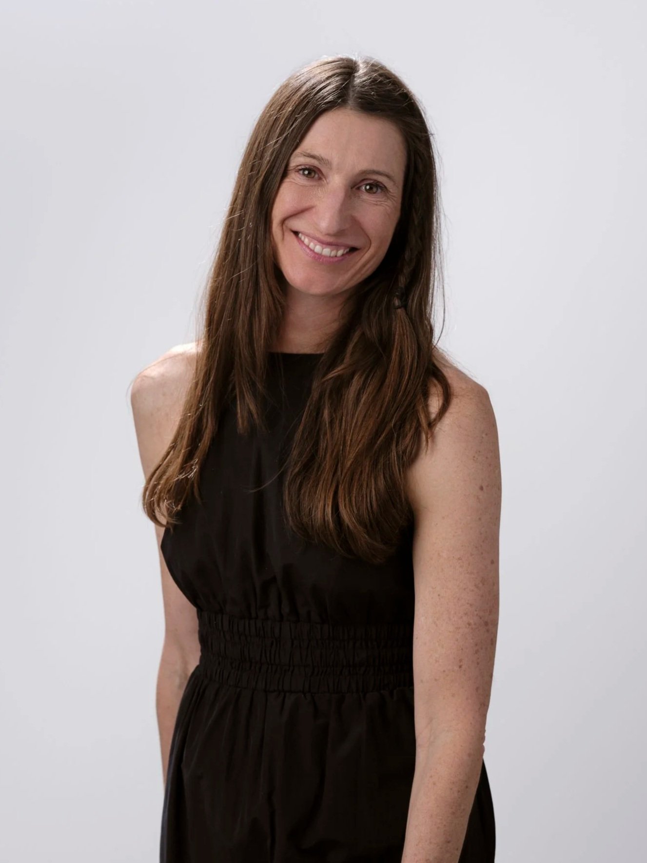 A woman with long brown hair, smiling, wearing a sleeveless black dress against a plain light-colored background.