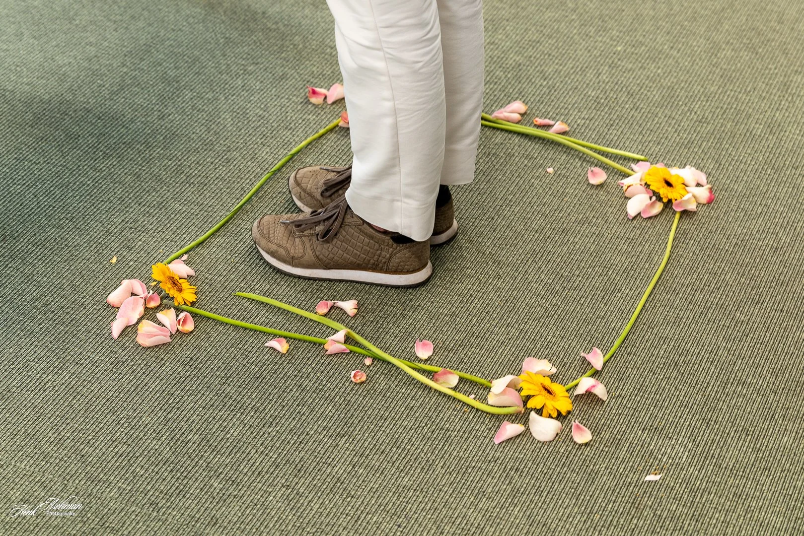Person standing inside a heart-shaped arrangement of flower stems, yellow and pink flowers, with scattered flower petals on a green carpeted floor.