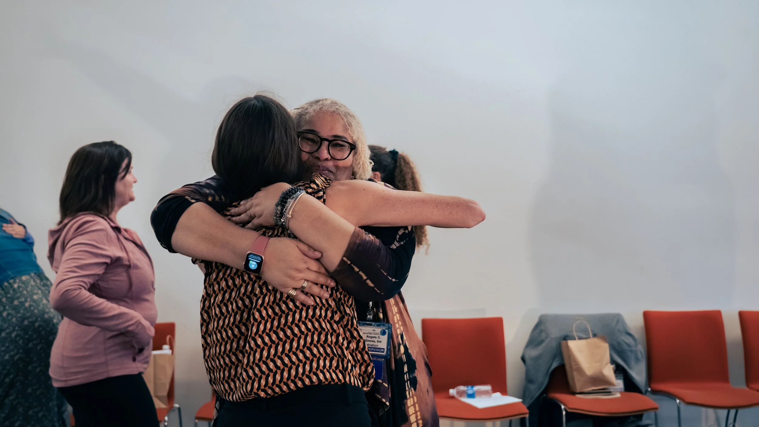 Two women embrace each other in a hug at an indoor event, with others standing nearby and an empty row of orange chairs in the background.