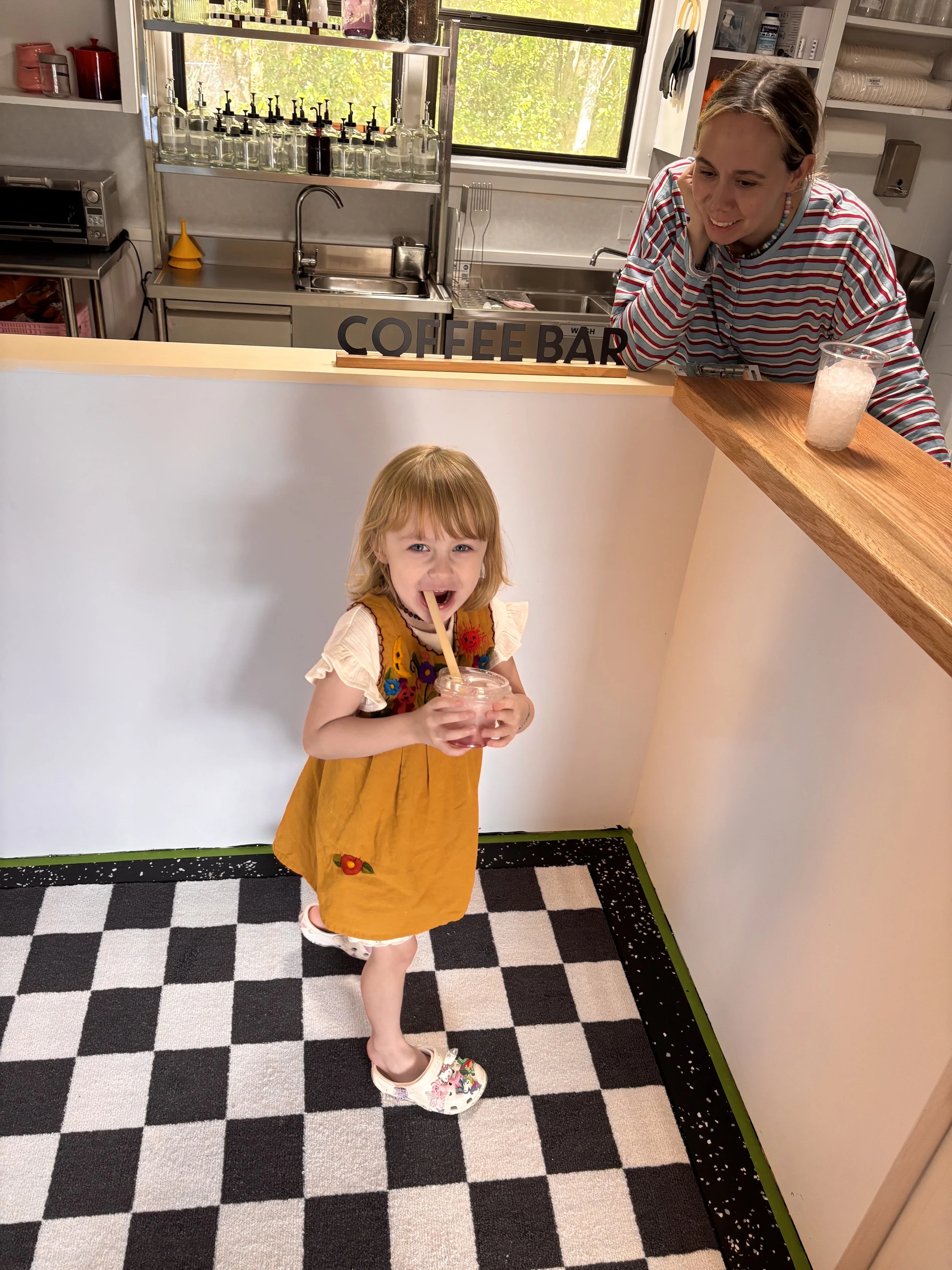 A young girl in a mustard yellow dress with colorful embroidery is standing on a black and white checkered floor, sipping a pink beverage through a straw. Behind her is a woman leaning over a counter at a coffee bar, smiling while talking on the phon