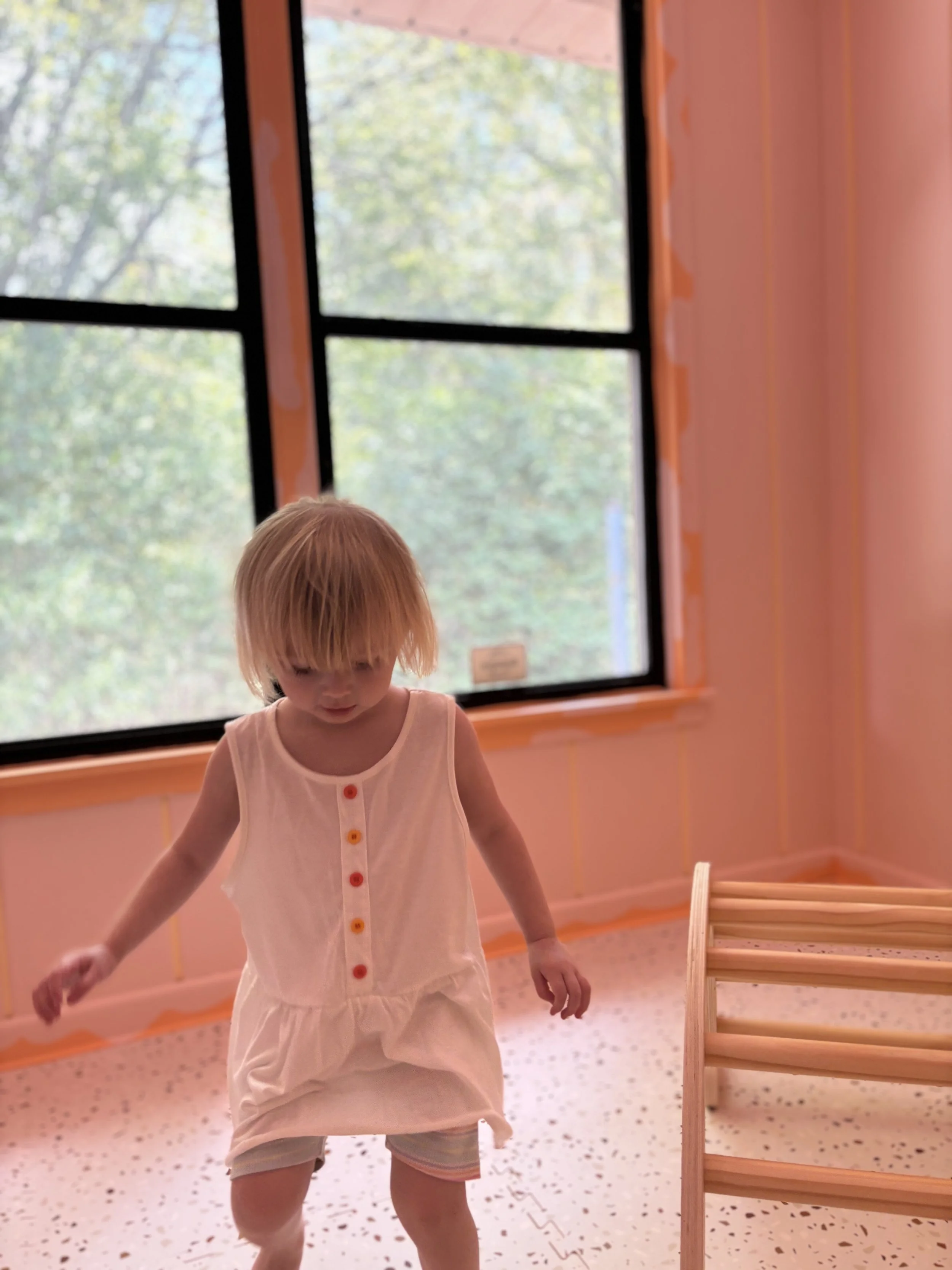A young girl with short blonde hair wearing a sleeveless, white dress with colorful buttons, walking in a room with peach-colored walls and a large window showing blurry trees outside.