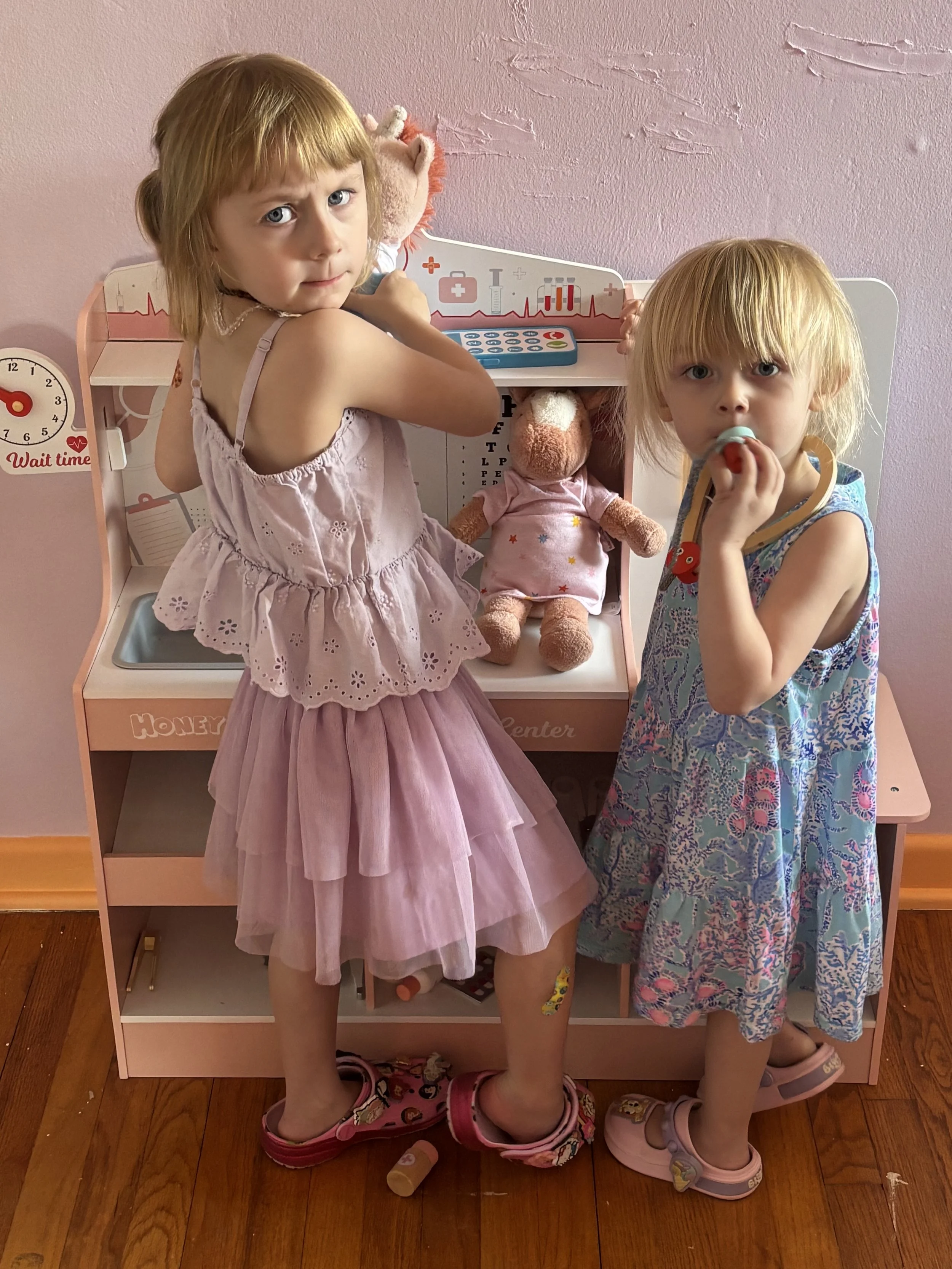 Two young girls with blonde hair playing at a pretend doctor or hospital play set. One girl is wearing a pink dress and pink shoes, and the other is wearing a colorful dress and white sandals. They are engaged with the toy hospital, which has a stuff