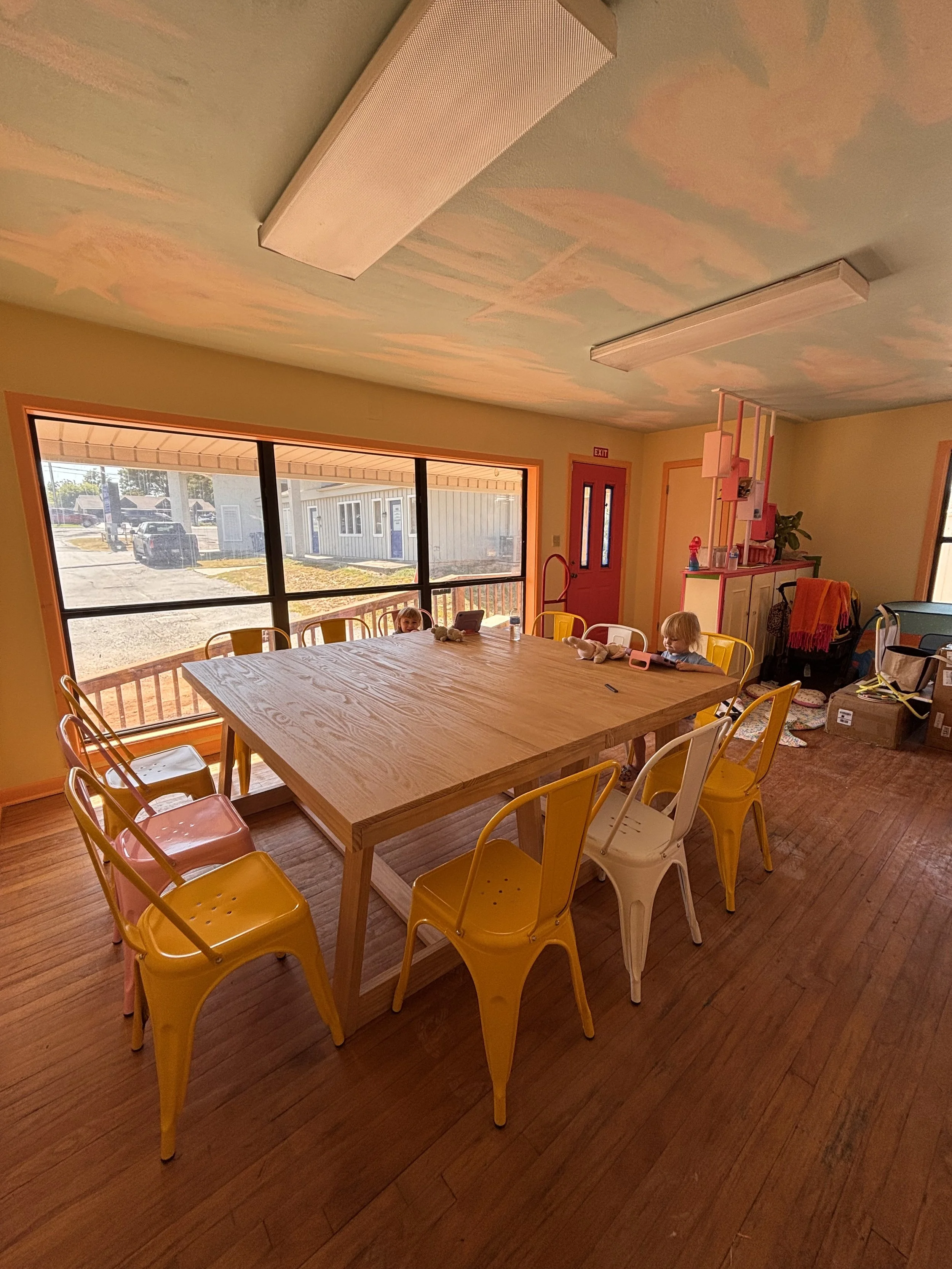 Inside a room with a large wooden table surrounded by yellow and white chairs, with children on the chairs. The room has large windows showing an outside view of parked cars and buildings, and the ceiling has painted clouds. There are various items l