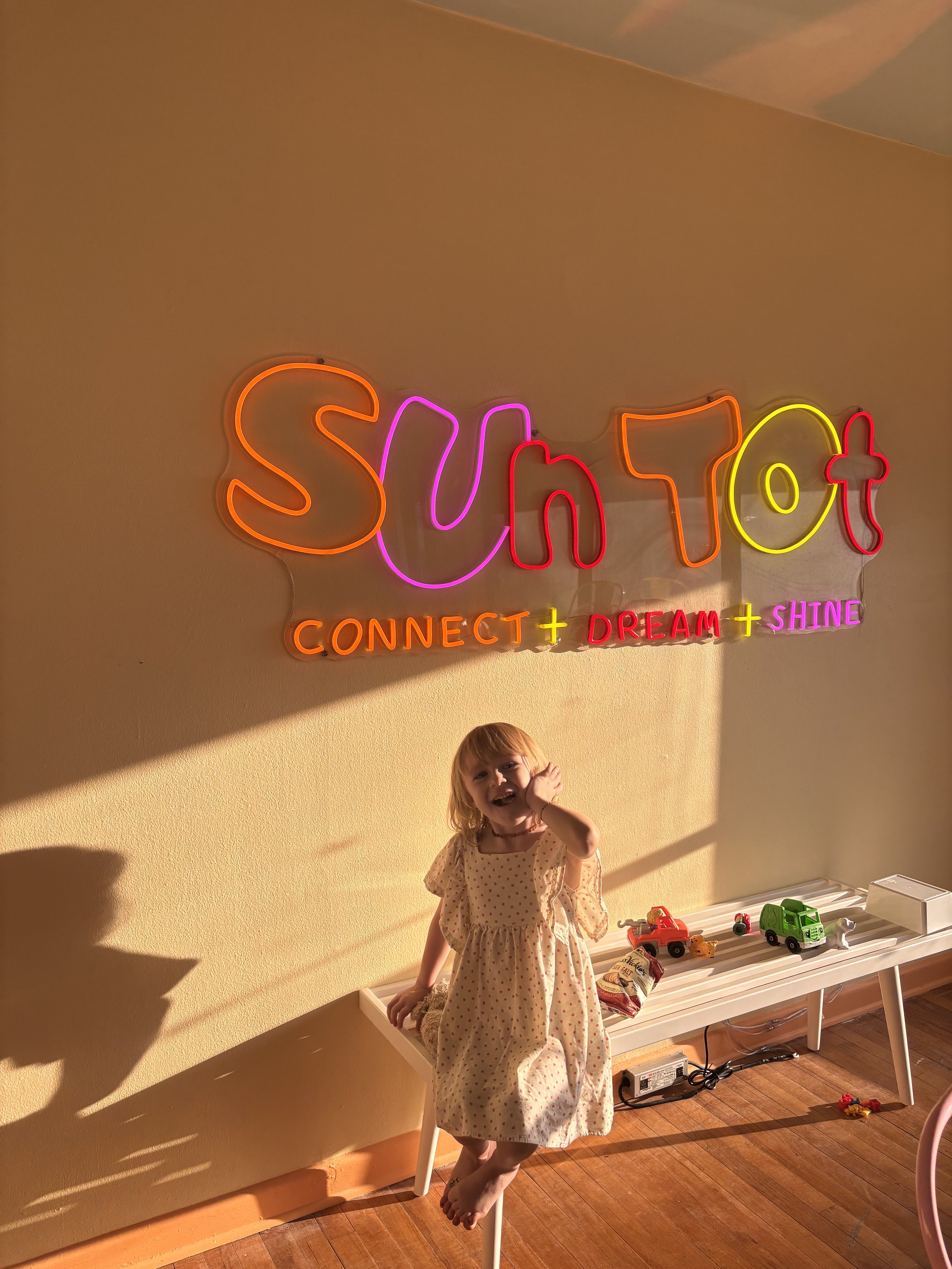 Neon sign that says "Sun Tot" with the words "Connect," "Dream," and "Shine" underneath. A young girl in a polka dot dress is sitting on a white bench below the sign, smiling and holding one foot with one hand. Toys are on the bench and the floor.