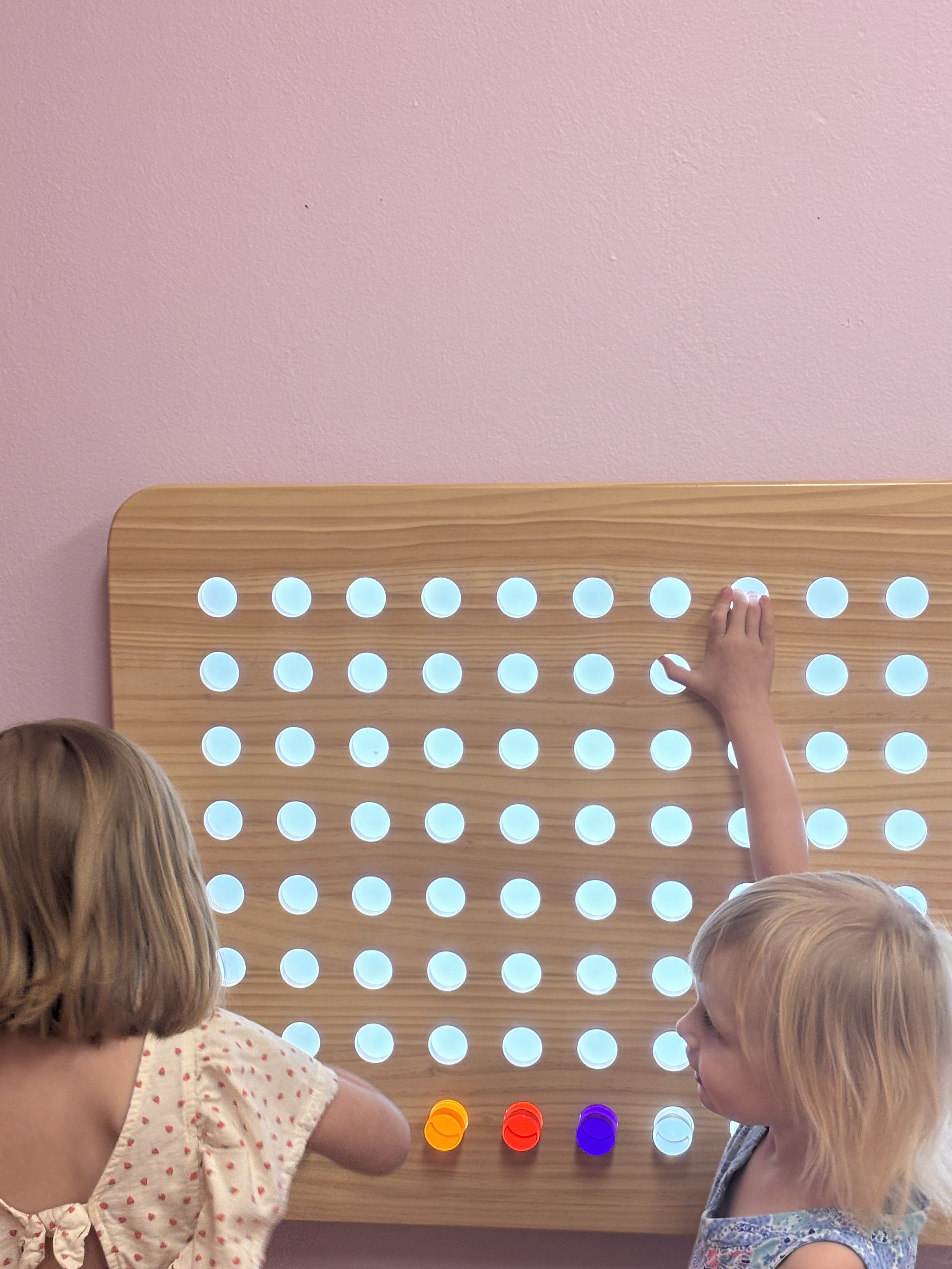 Children playing a game of Connect Four with falling colored discs on a wooden game board against a pink wall.
