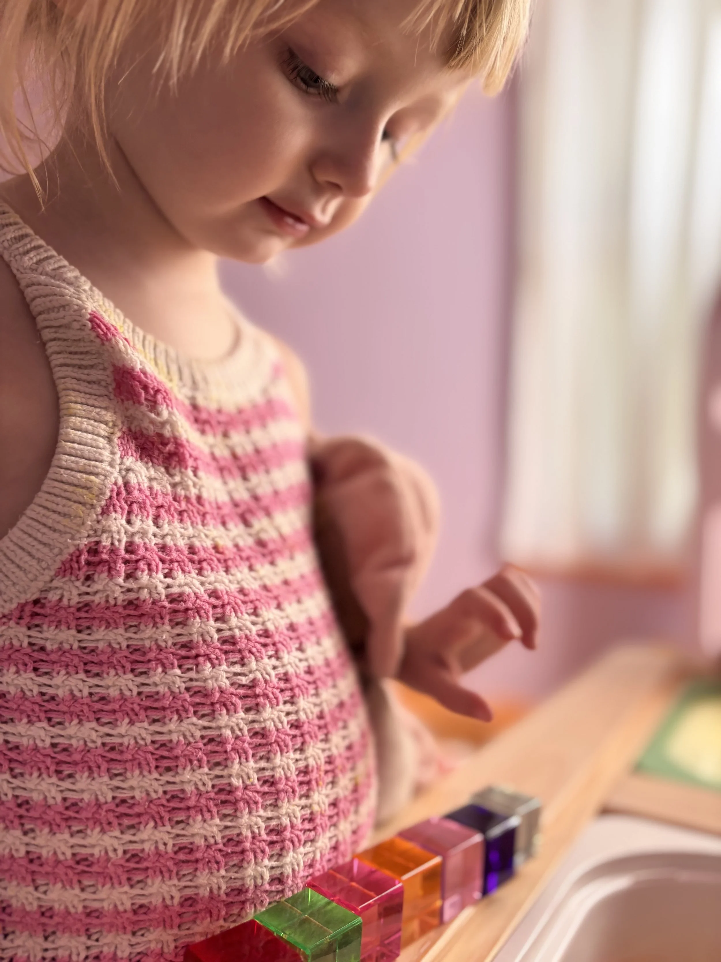 Close-up of a young girl with blonde hair and a pink, white, and beige striped knitted sweater, looking down at a row of colorful her in a play area, with a pink wall and window with curtains in the background.