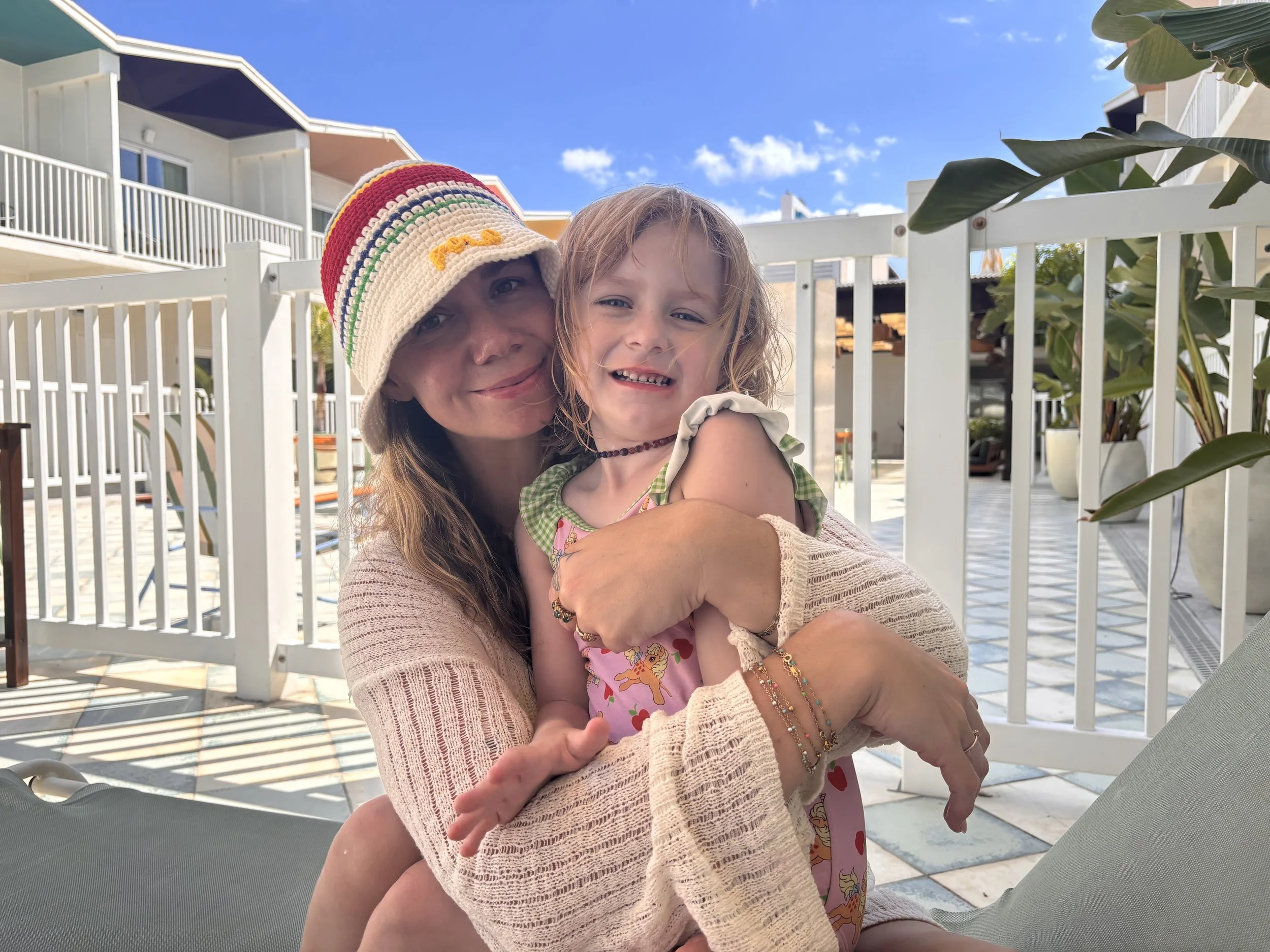 A woman wearing a colorful striped knit hat and a beige knitted cardigan holds a smiling young girl with light brown, curly hair, wearing a pink dress with cartoon characters and jewelry on her arms and neck. They are outdoors on a sunny day, in front of a white fence, with buildings and trees visible in the background.