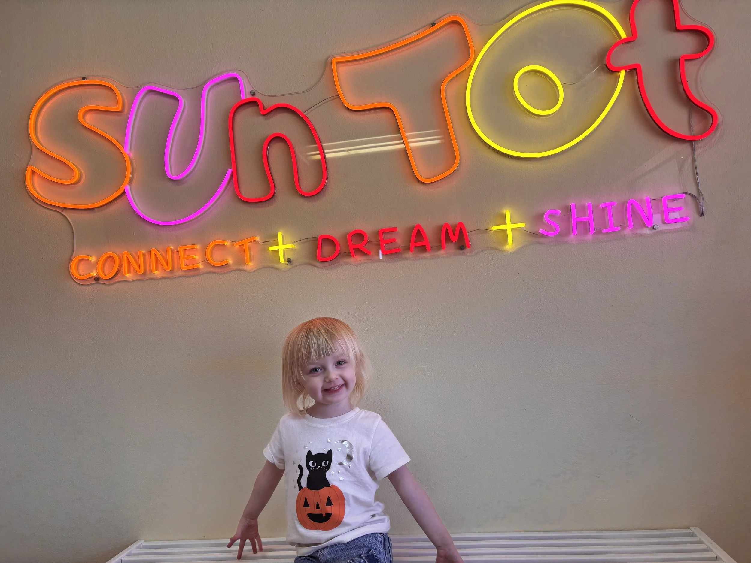 A smiling young girl with blonde hair and blue eyes standing in front of a beige wall with a colorful neon sign that reads 'SHINE' and 'CONNECT DARE + SHINE'. The girl is wearing a white T-shirt with a black cat and pumpkin design and blue jeans.