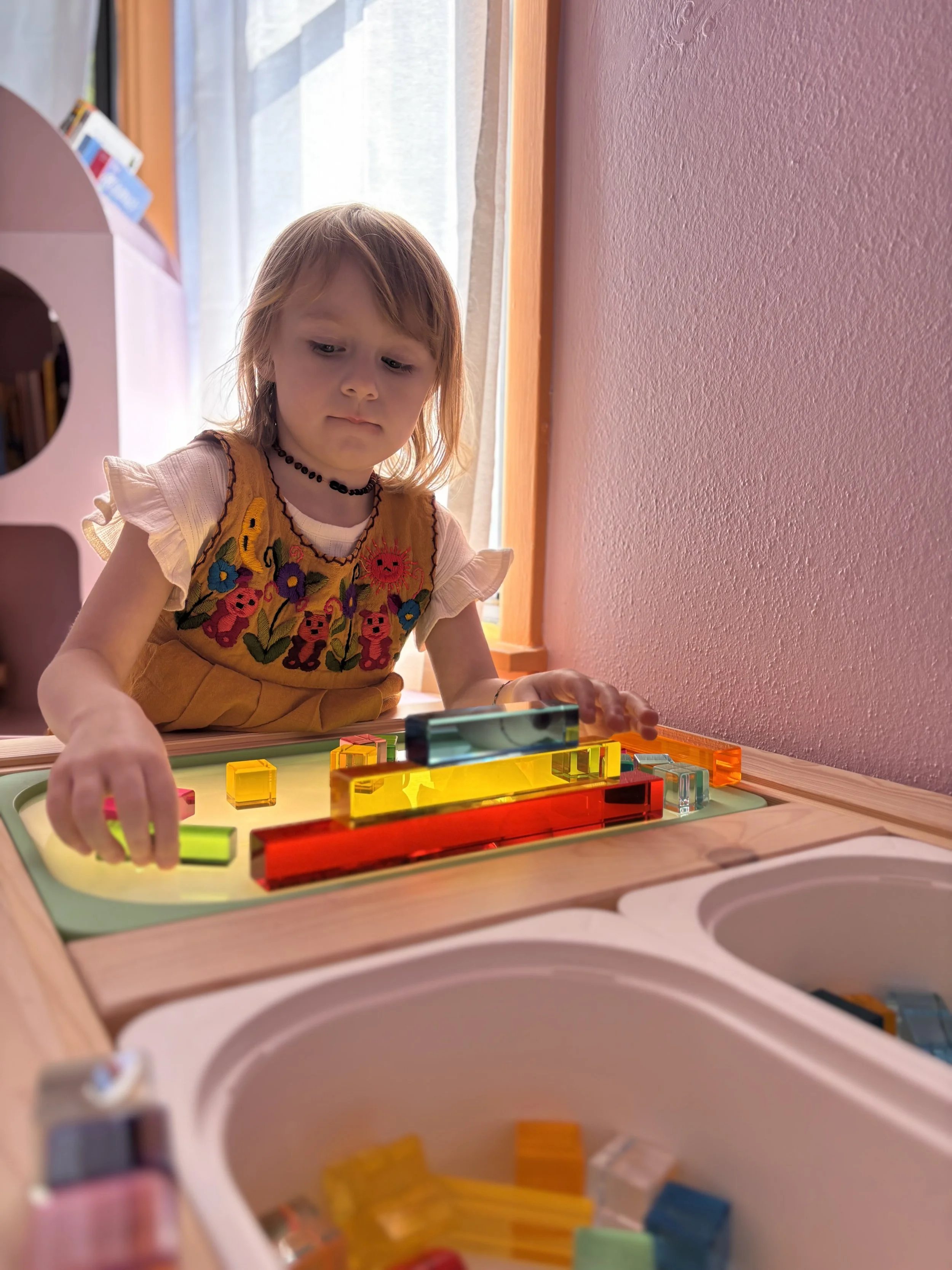 A young girl with reddish-brown hair, wearing a yellow dress with colorful embroidery, playing with colorful translucent building blocks at a wooden table near a pink wall and a window.