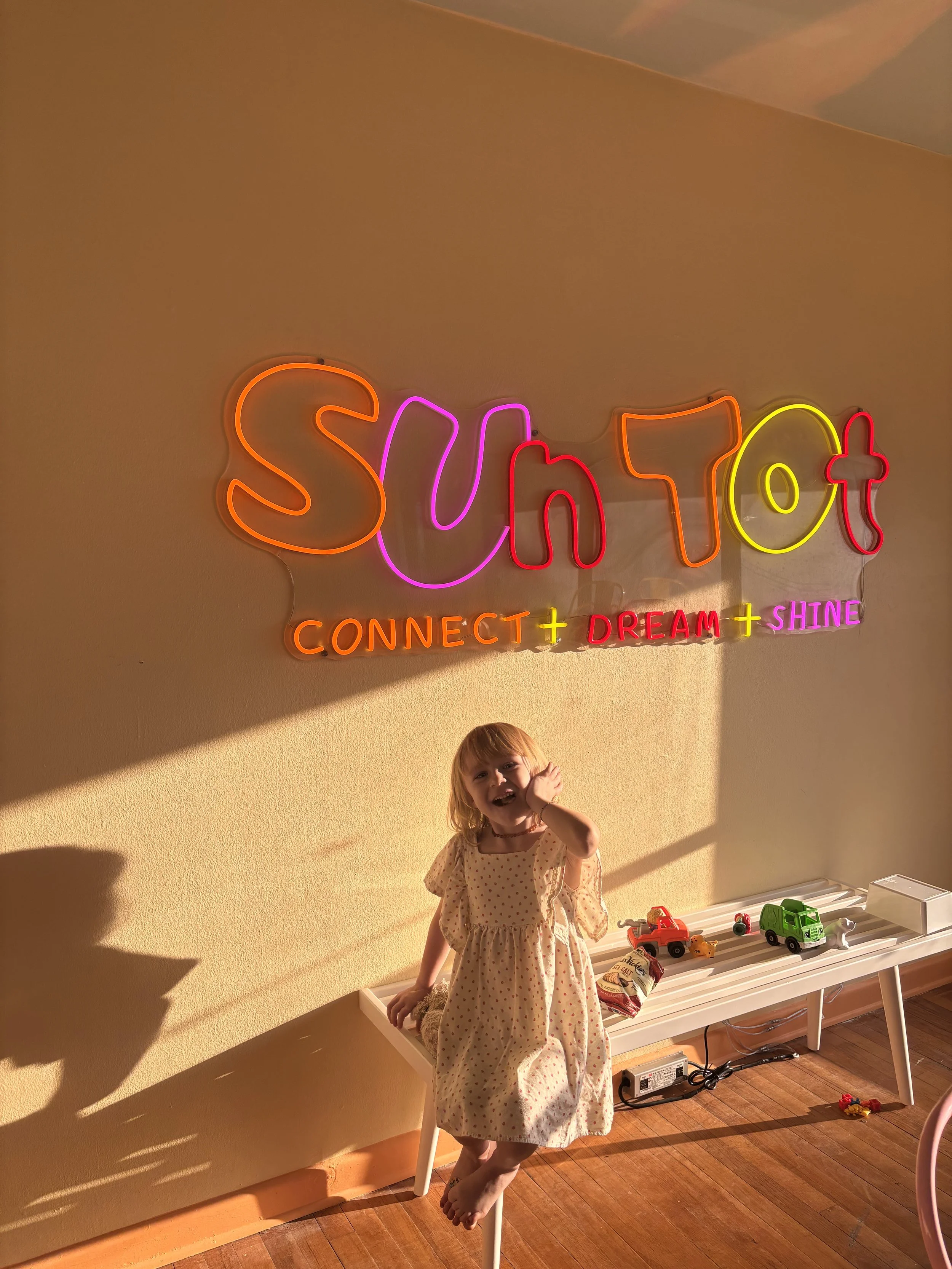 A young girl in a polka dot dress is standing on a white table, smiling and touching her face with her left hand. Behind her, there is a neon sign on the wall that reads 'SUSHI' in colorful letters and 'CONNECT + DREAM + SHINE' below it. The room has