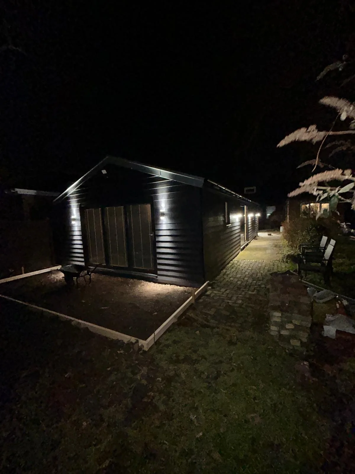 Nighttime view of a dark-colored house with exterior lighting along the side, a paved walkway, benches, and a small garden bed with a wooden border.