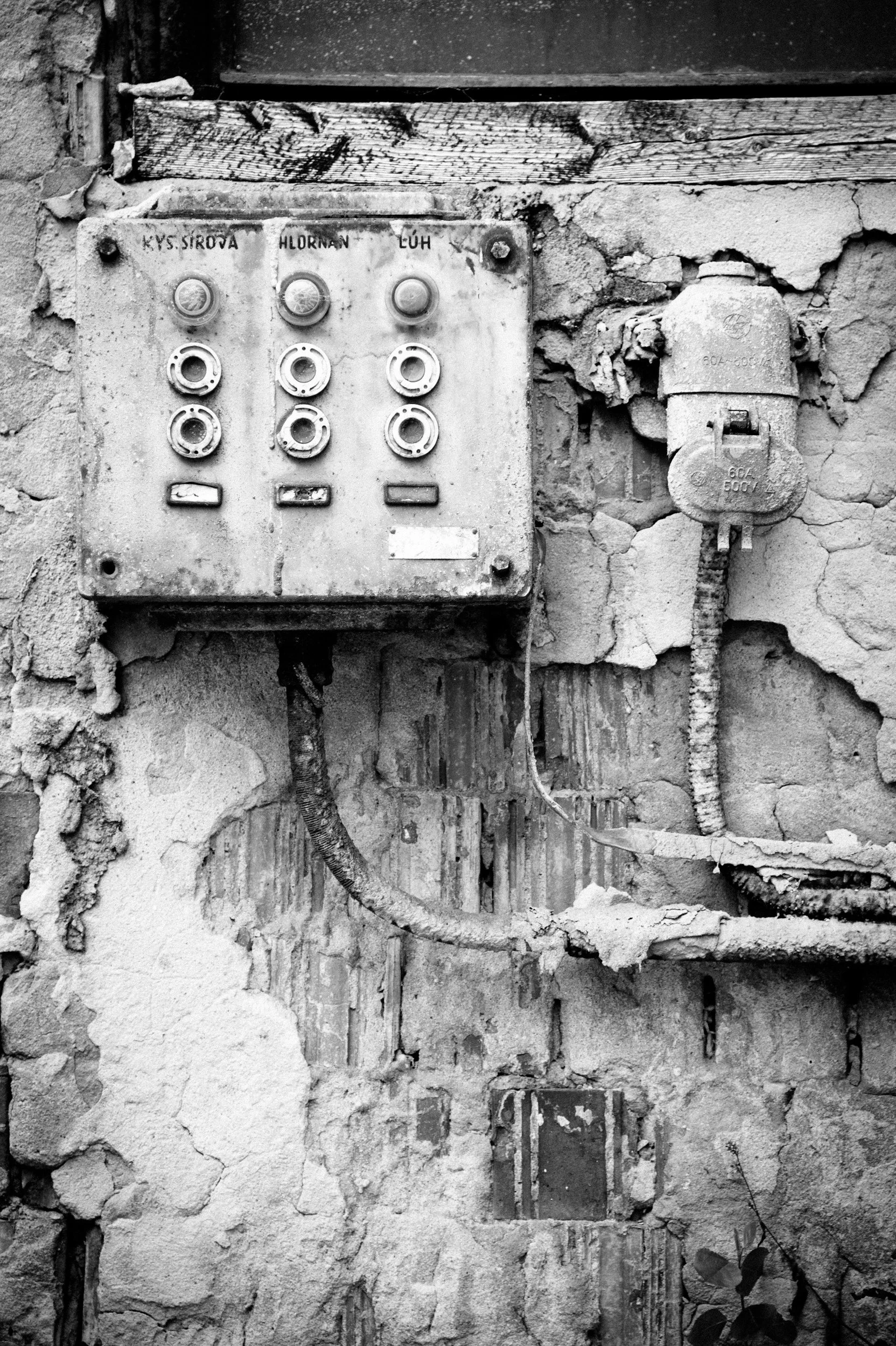 Black and white photo of an old, weathered electrical panel with several disconnected sockets and a plugged-in cord, mounted on a cracked wall with peeling plaster and exposed bricks.