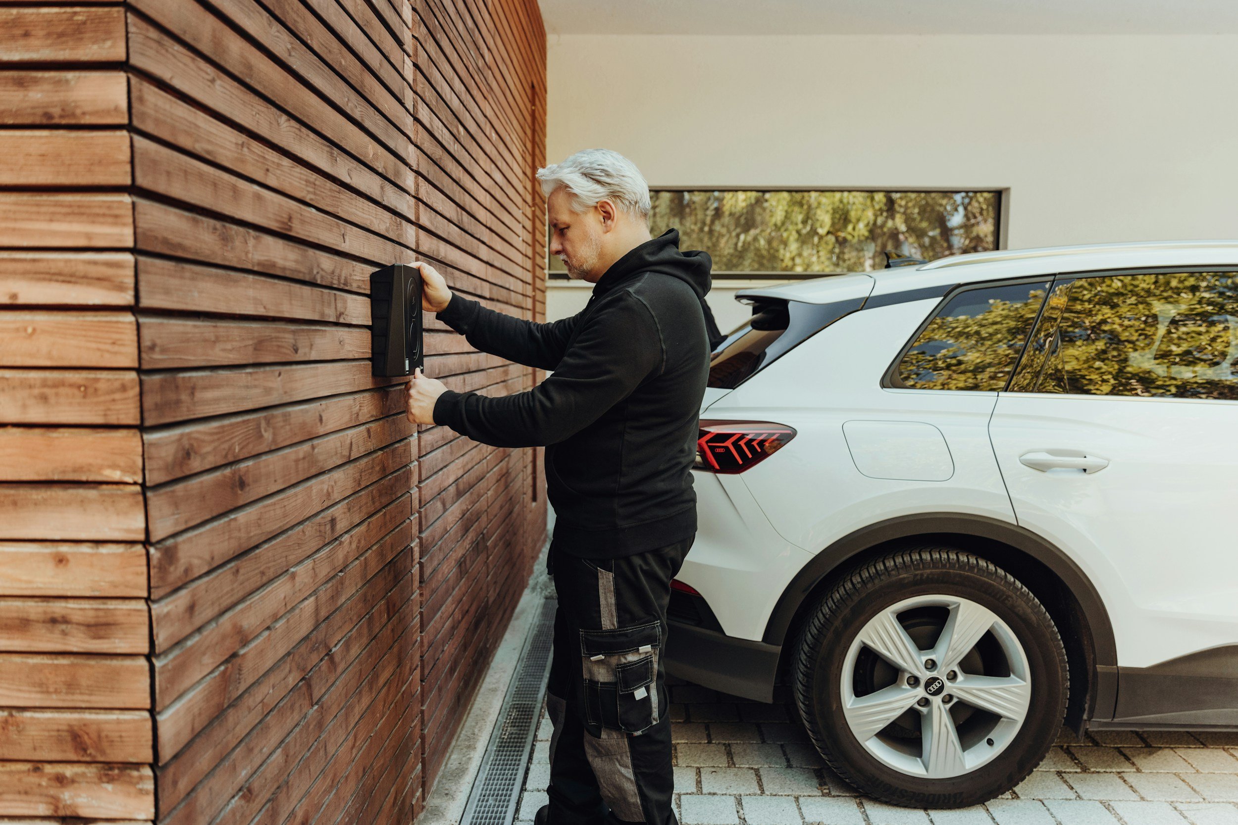 A man with gray hair and a black hoodie downloading a package from a mailbox attached to a wooden wall outside a building. A white SUV is parked nearby, reflecting trees in its windows.
