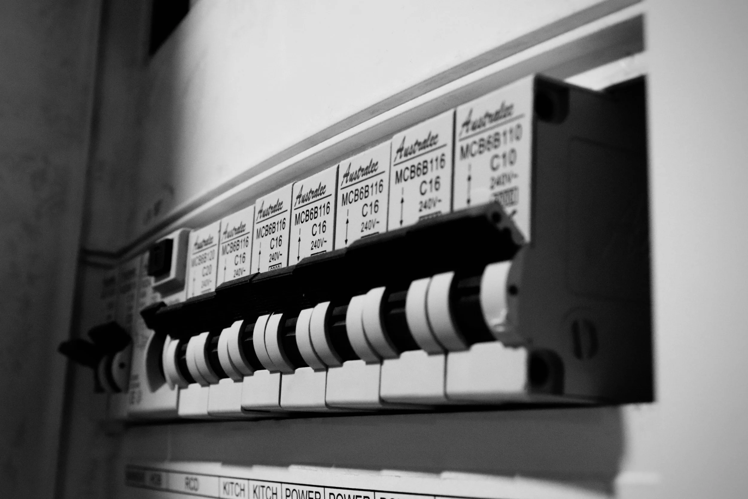 Close-up of a circuit breaker panel with labeled switches, some of which are switched on. The labels include 'KITCH POWER' and others with codes.