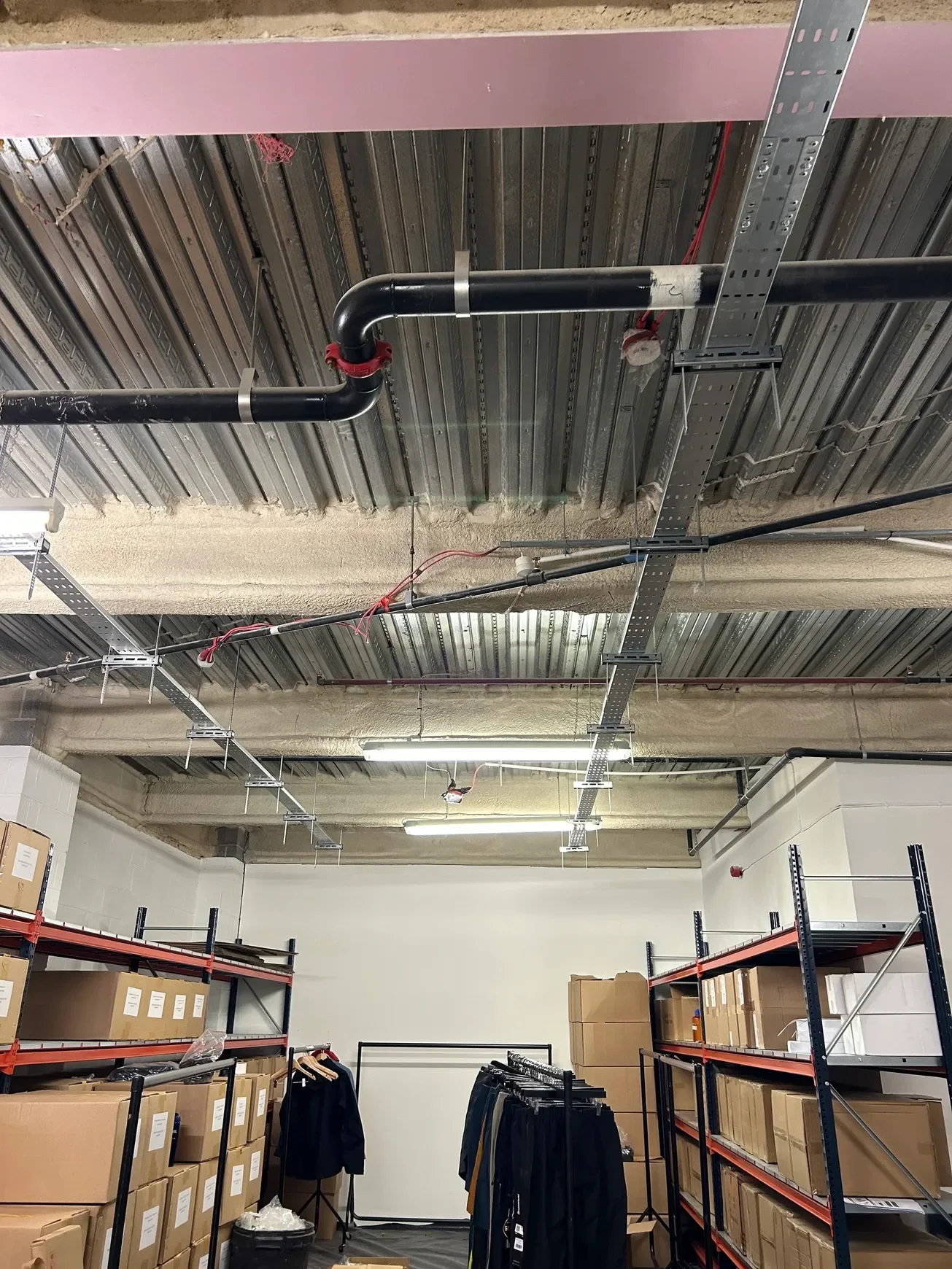 Warehouse with unfinished ceiling showing pipes and wiring, metal shelving units filled with boxes and clothing racks with hanging clothes.