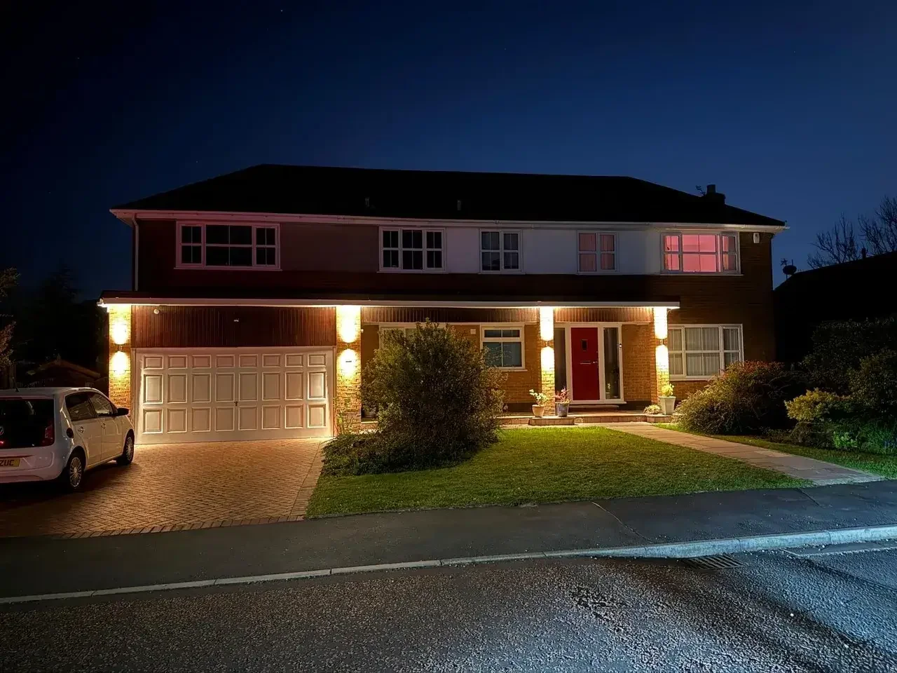 Two-story suburban house illuminated at dusk, with a double garage, red front door, landscaped lawn, and parked car.
