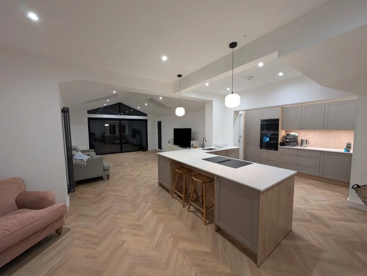 Modern open-plan kitchen and living room with a large island, pendant lights, gray cabinetry, herringbone flooring, and glass doors