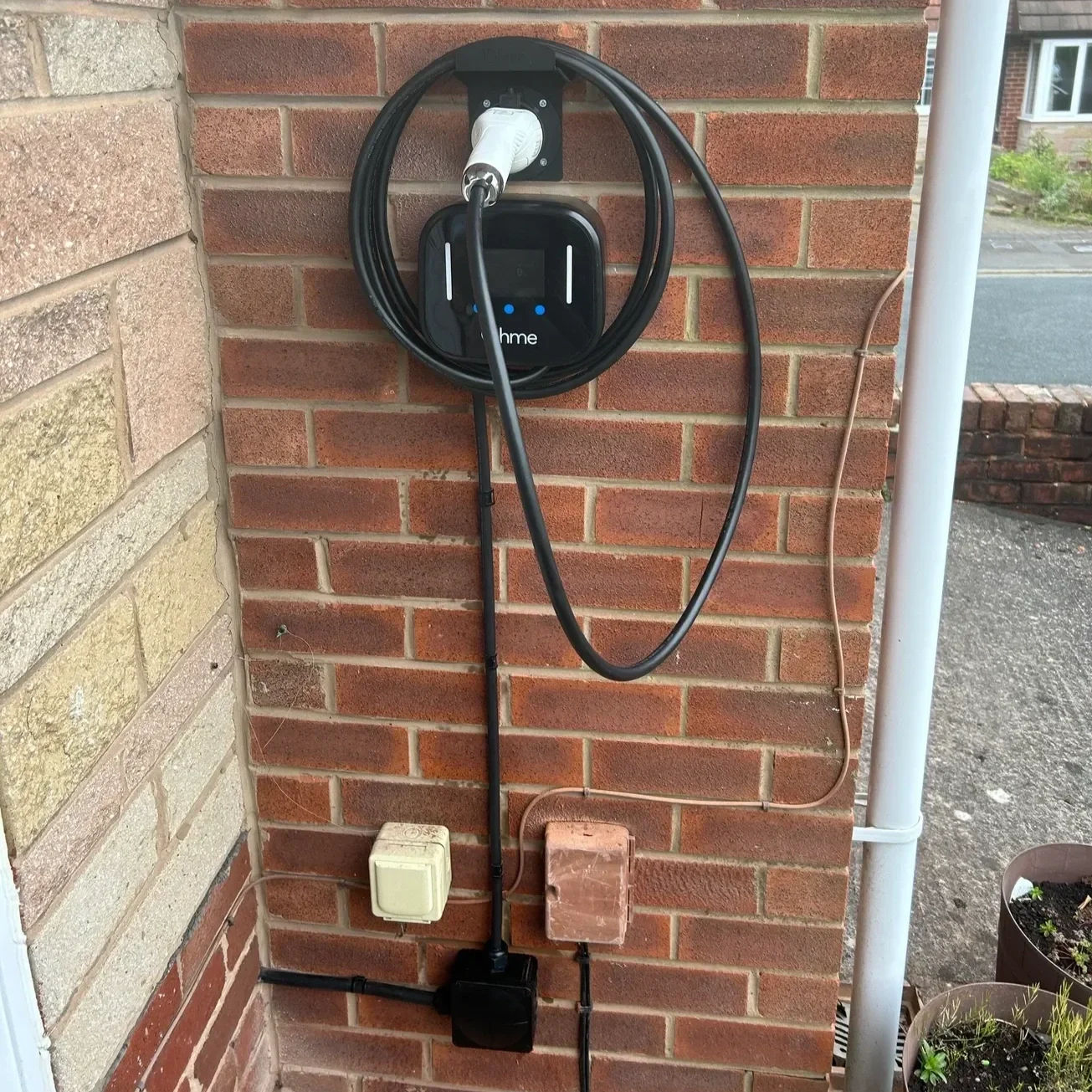 Electric vehicle charging station mounted on a brick wall, with a charging cable and connector attached, located outdoors near a street.