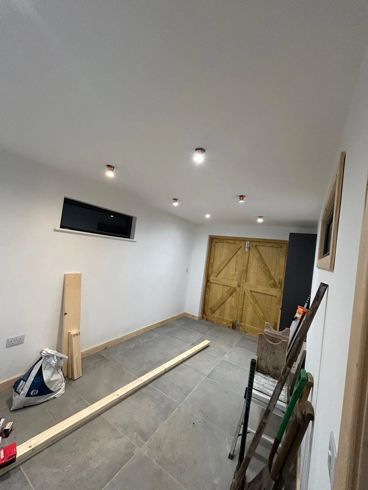 A room under construction with gray tiled flooring, four ceiling lights, a small horizontal window, and a wooden double door. Construction materials and tools are scattered on the floor and leaning against the wall.