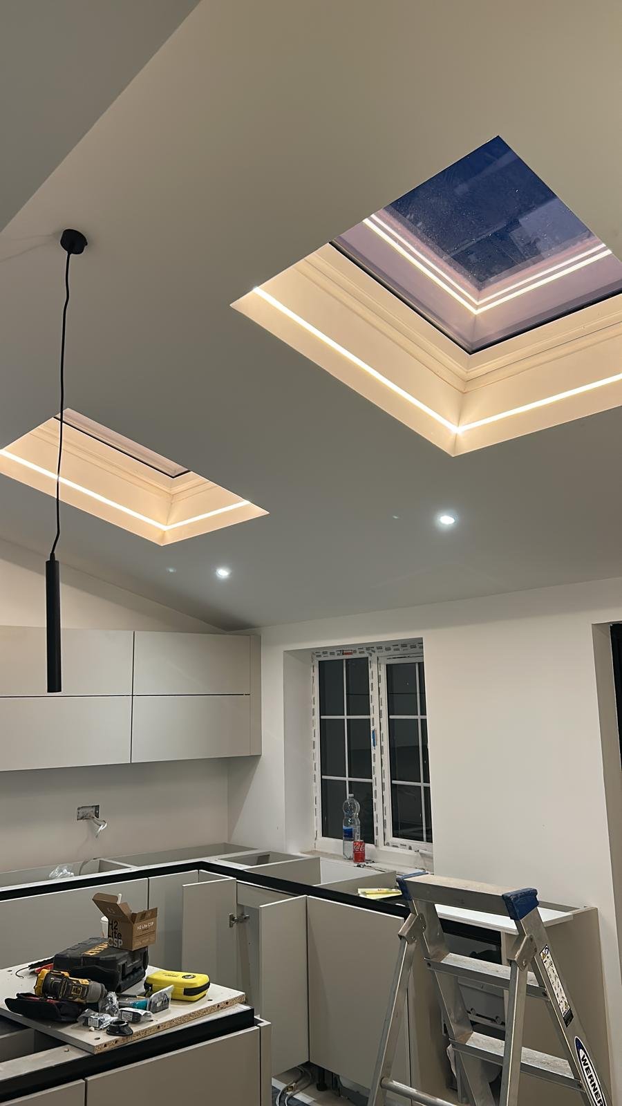 Kitchen under construction with white cabinets, a window, construction tools, and a ceiling with recessed lighting and skylights.