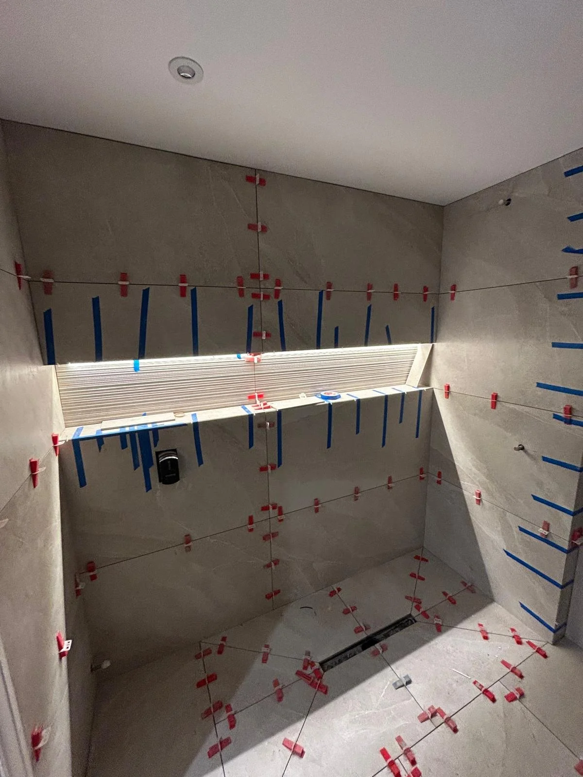 Bathroom under construction with tiles being installed, red and blue spacers, and a louvered window.