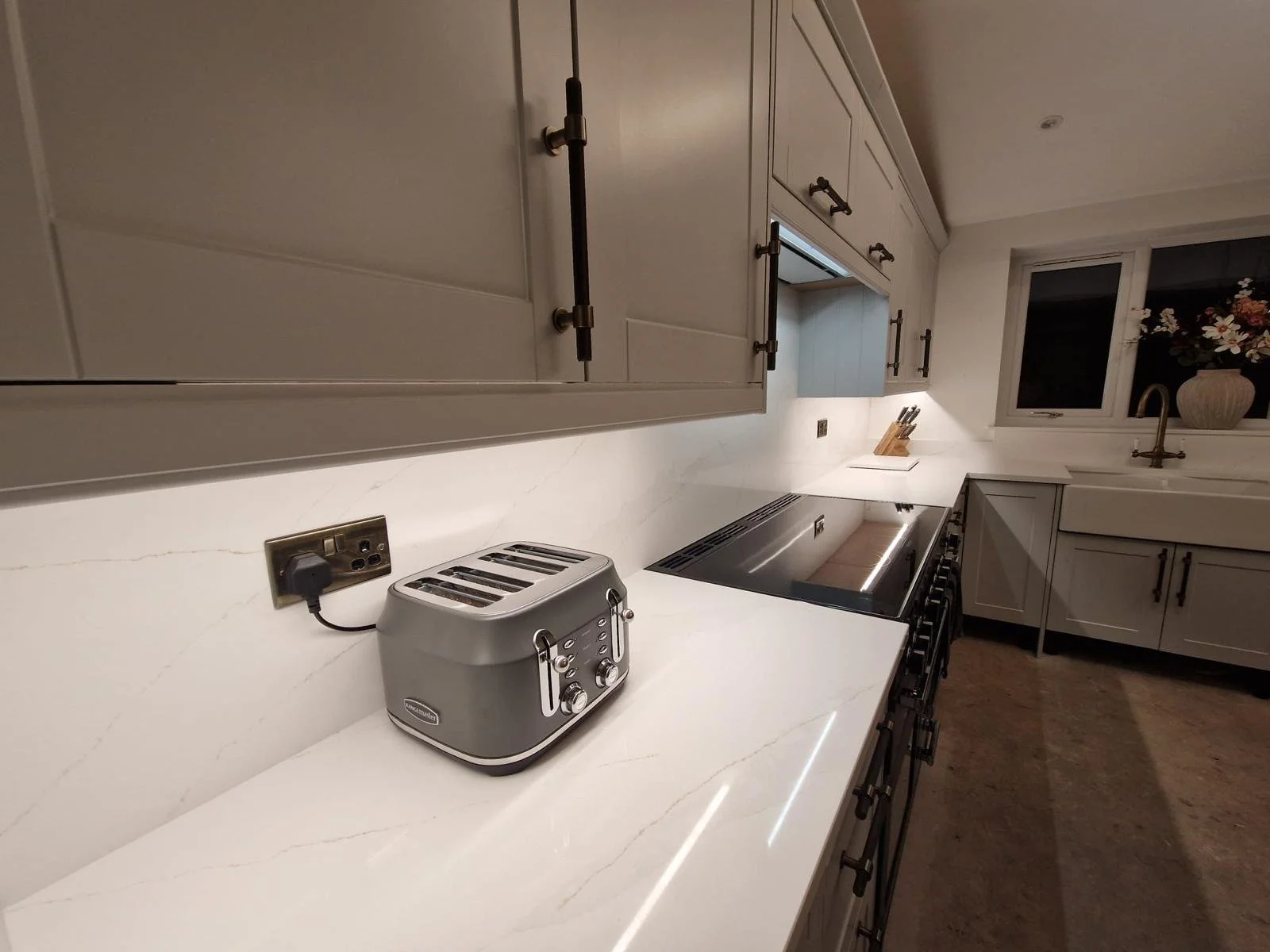 Kitchen with white marble countertops, beige cabinets, black handles, a toaster, and a window above the sink with a vase of flowers.