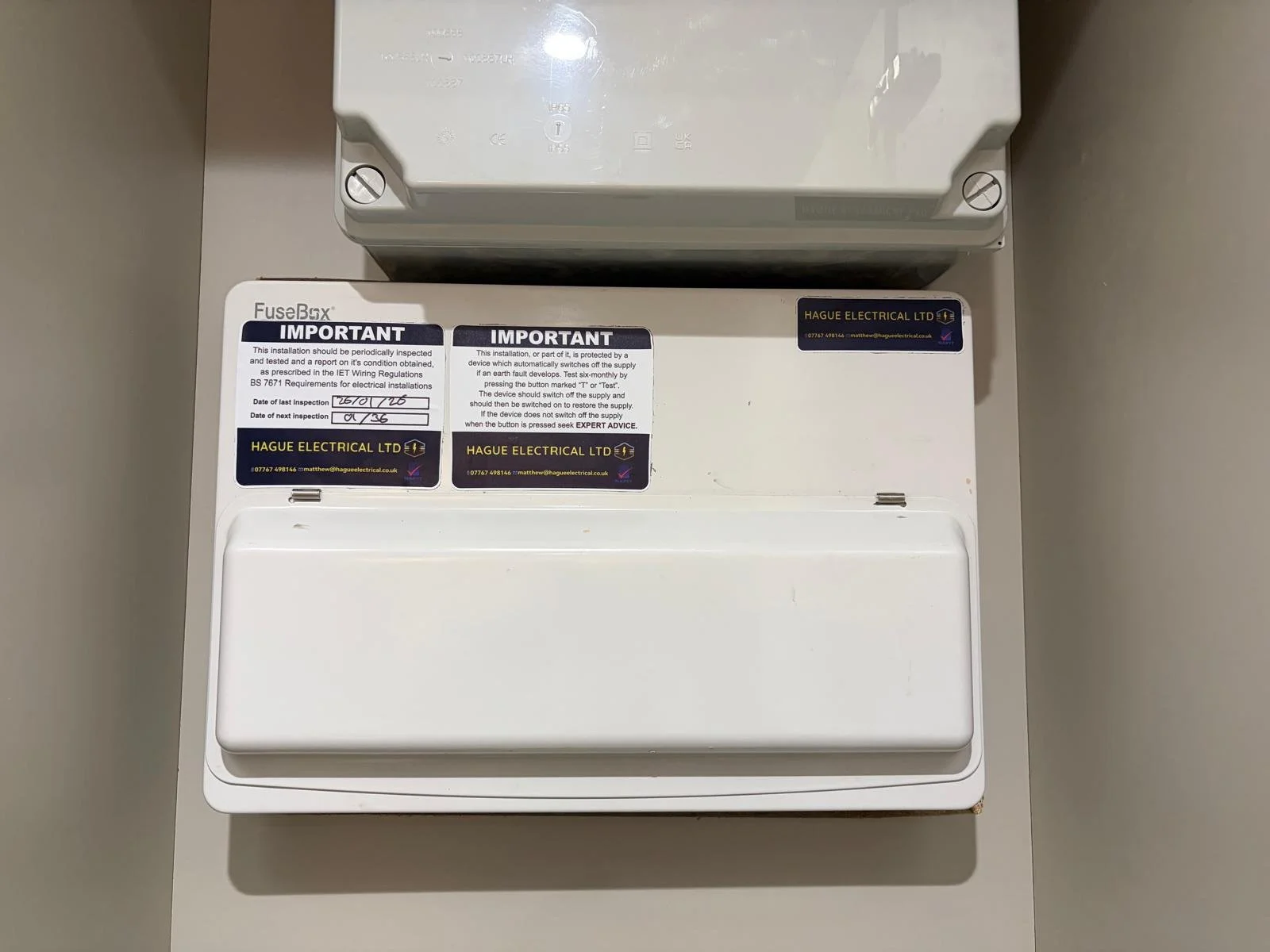 Close-up view of an electrical fuse box inside a white enclosure with maintenance stickers from Hague Electrical LTD indicating inspection dates and details.