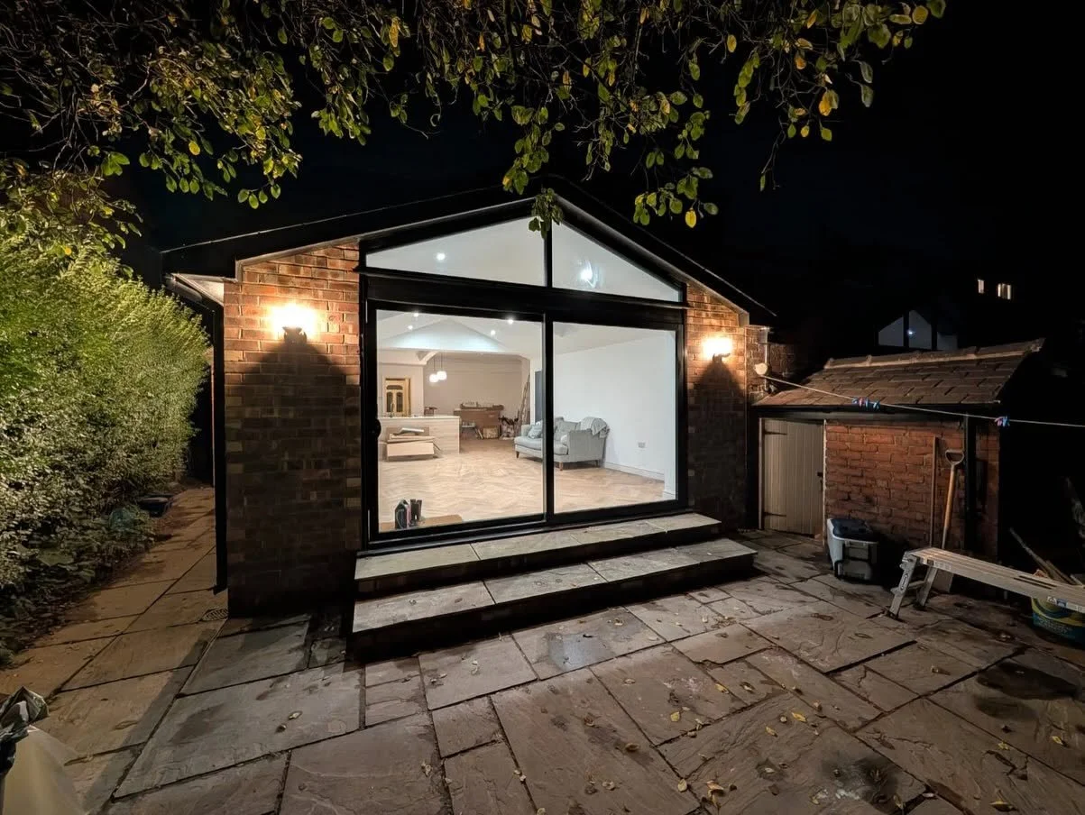 Nighttime view of a brick home extension with large glass doors opening onto a paved patio and illuminated modern living space.