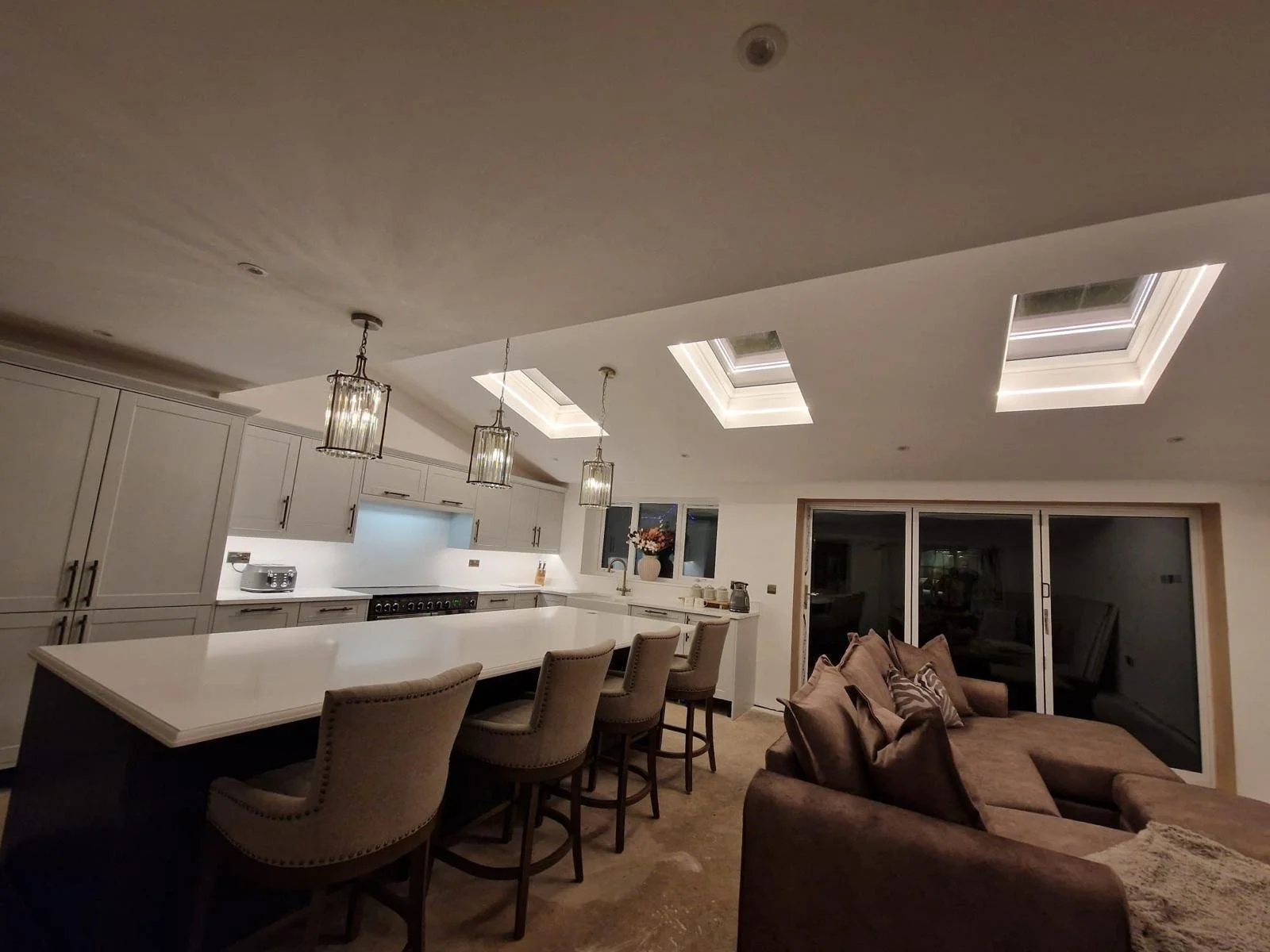 A modern kitchen with white cabinetry, a white central island with seating, pendant lighting, skylights in the ceiling, and a sliding glass door leading to a living room area with a brown sofa.
