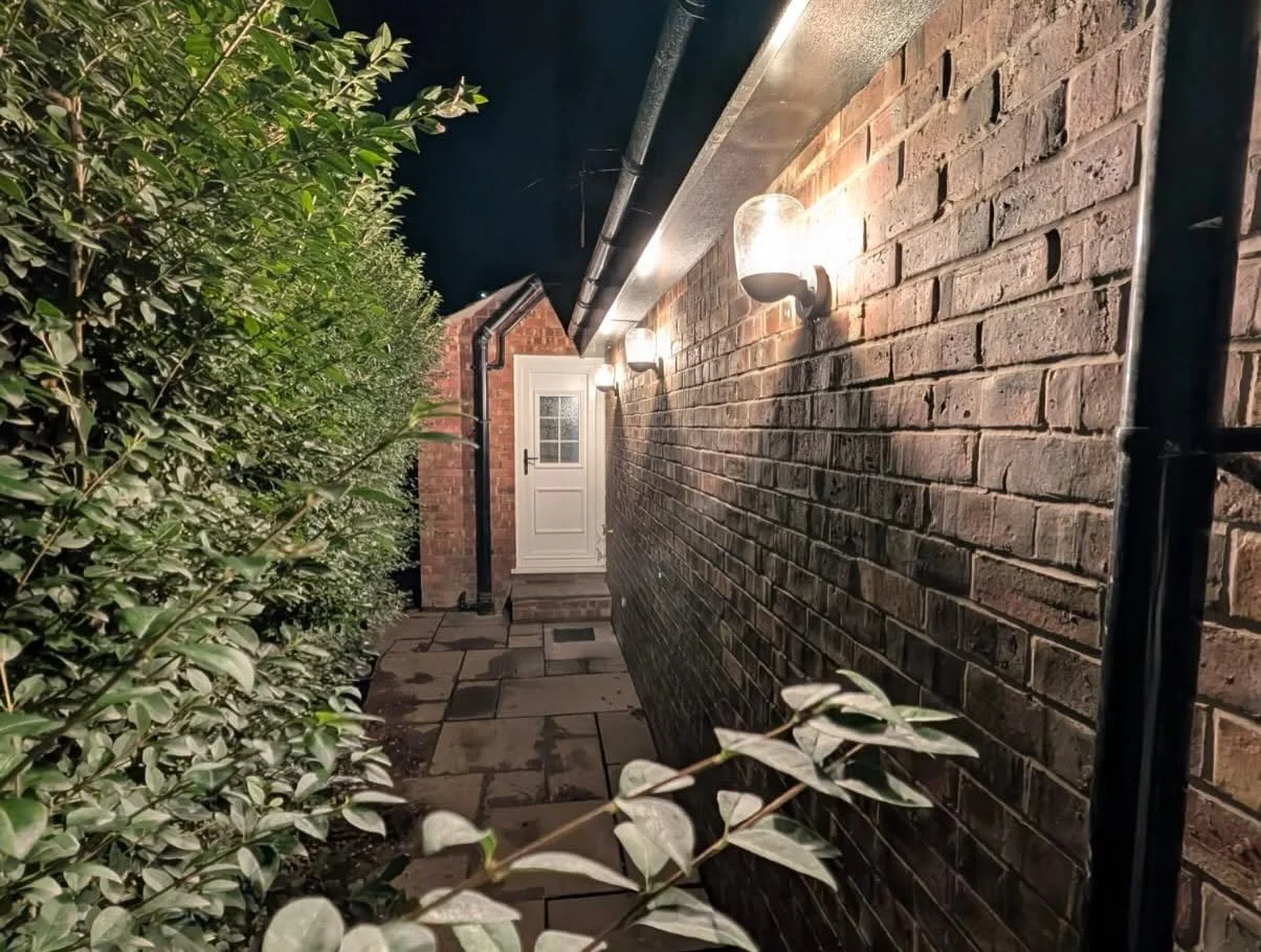 Nighttime view of a brick house side passage with illuminated wall lights, stone paving, greenery, and a white back door.