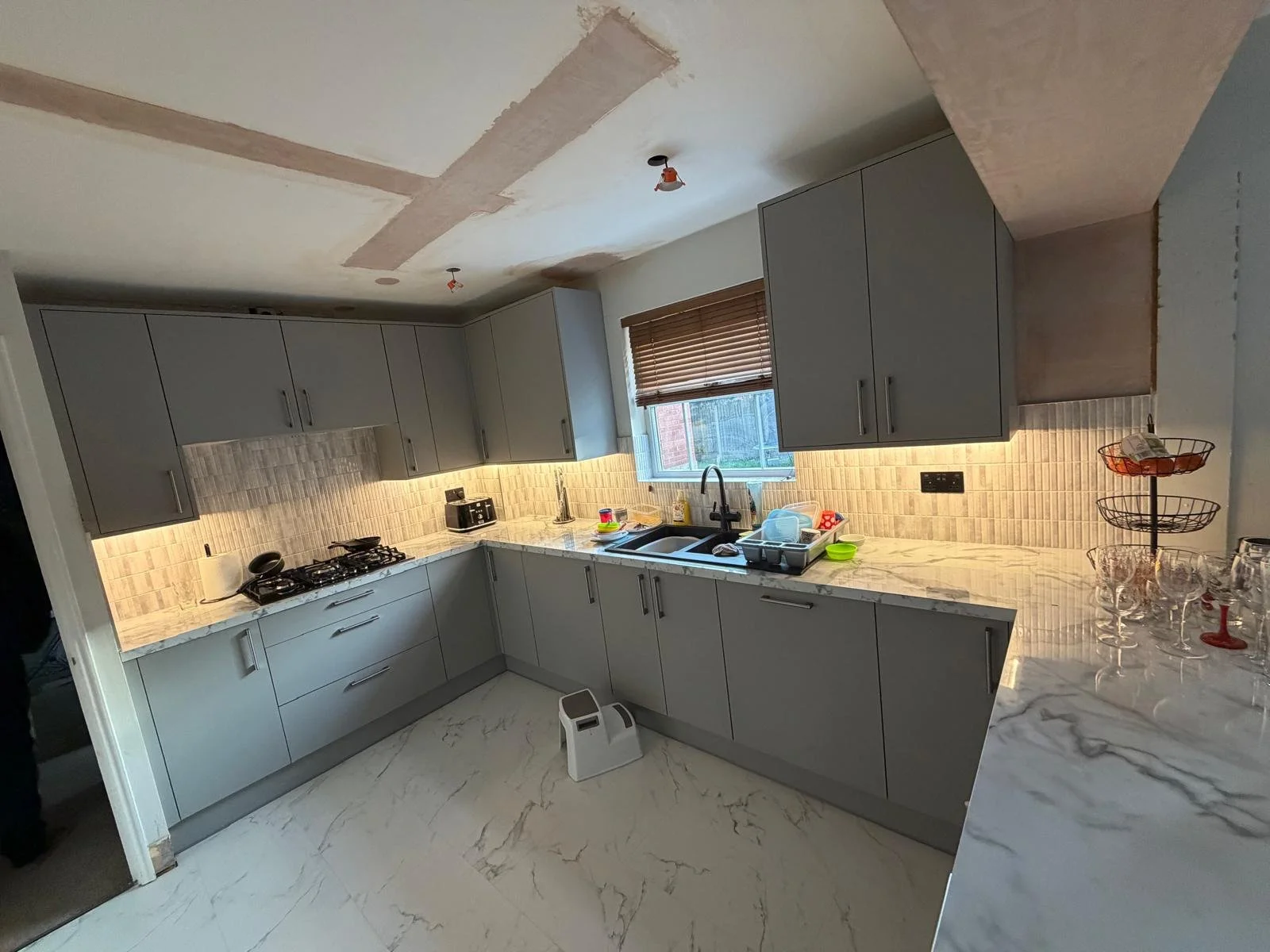 Modern kitchen with gray cabinets, marble countertops, and a beige tiled backsplash. Kitchen items include a stove, sink, various dishes, and glasses on the countertop near a window with wooden blinds.