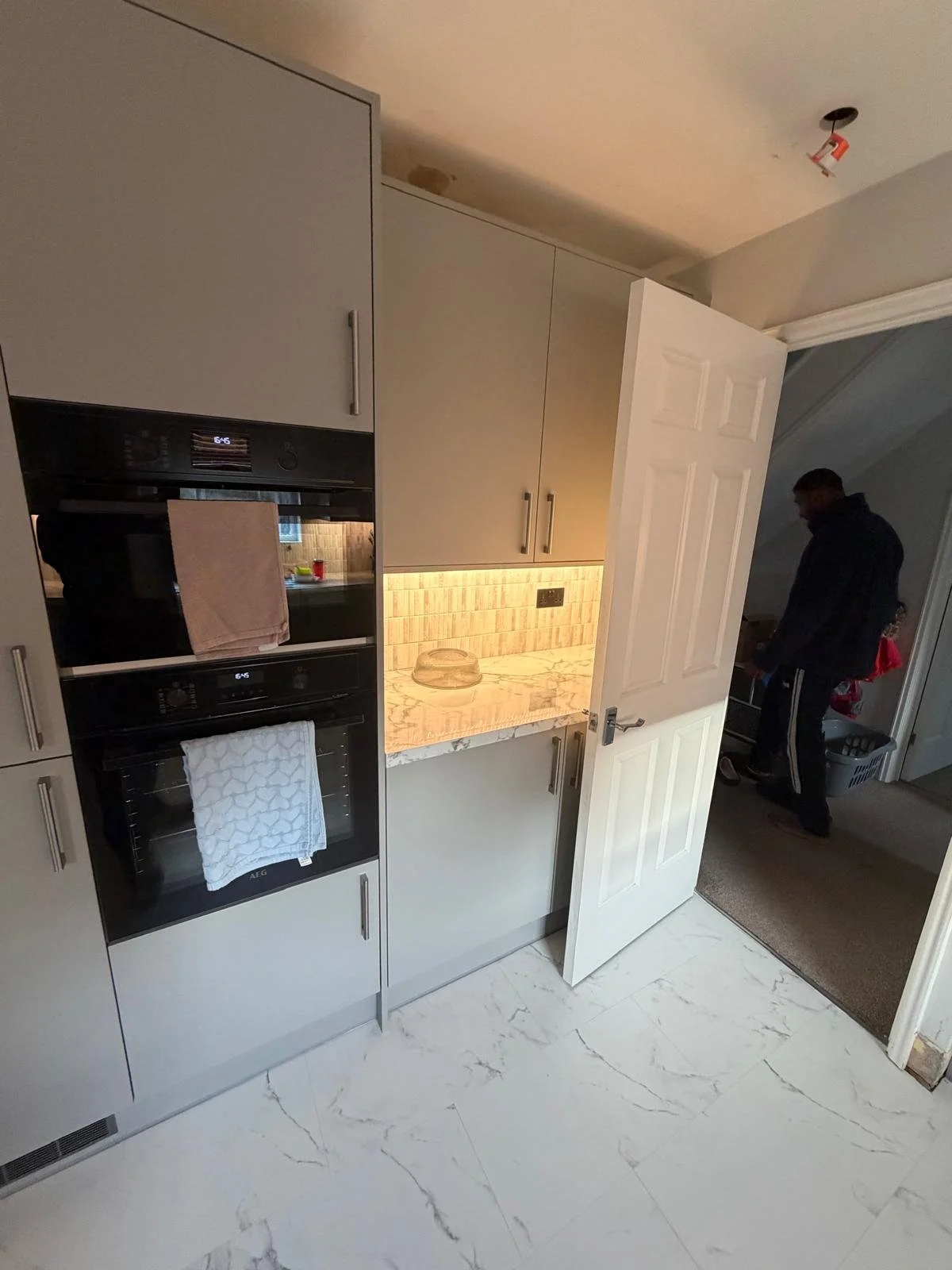 View of a modern kitchen with gray cabinets, a marble countertop, and a built-in oven. Towel hanging on the oven handle, and a glass bowl on the counter. Open door reveals a man in black clothing in an adjoining room.