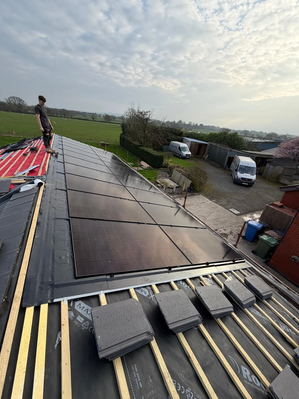 A person standing on a roof installing solar panels with a rural landscape in the background, including open fields, trees, and parked vans.