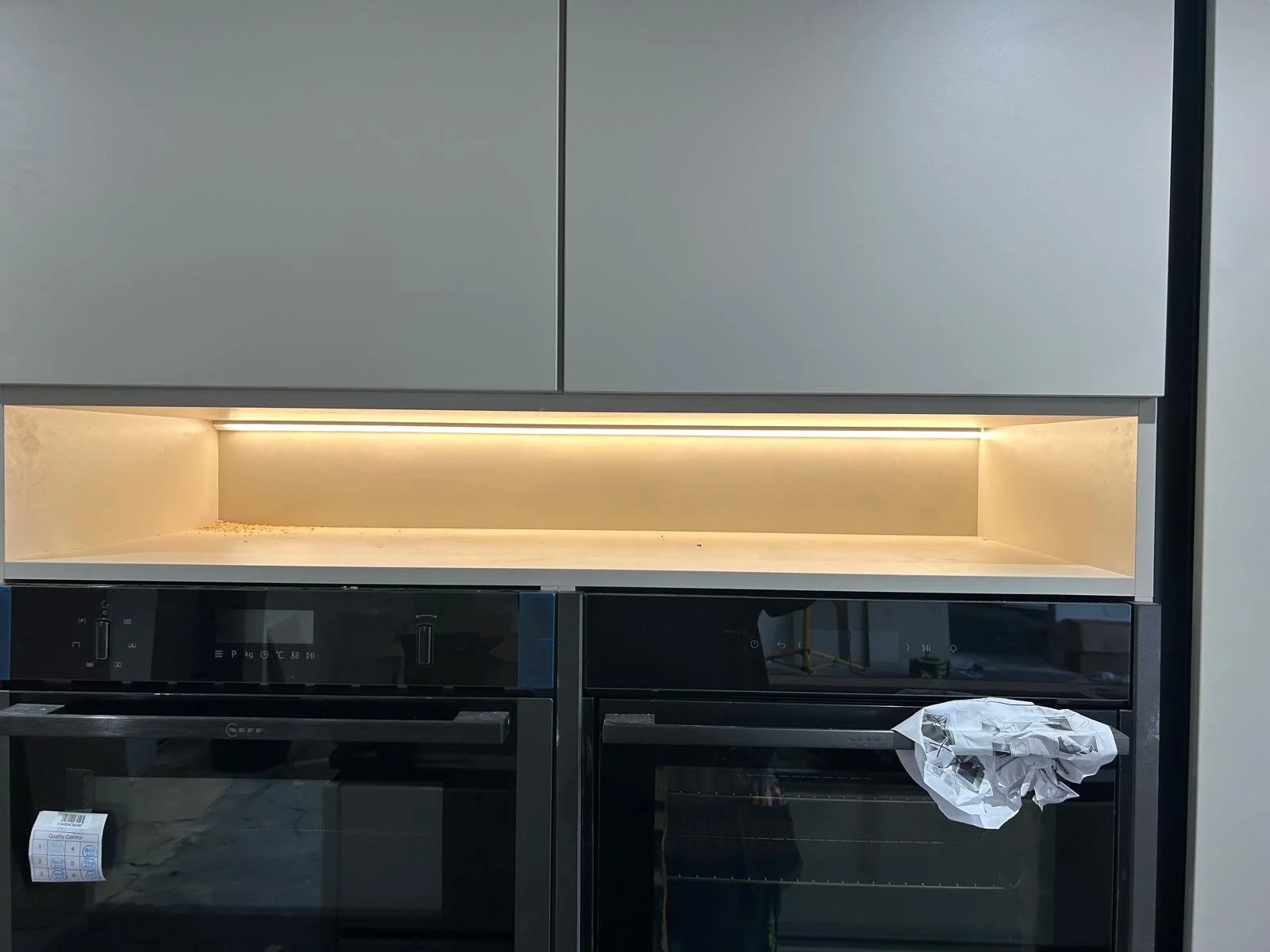 Empty kitchen cabinet with led lighting underneath, above built-in ovens, with a crumpled paper towel on the oven door handle.
