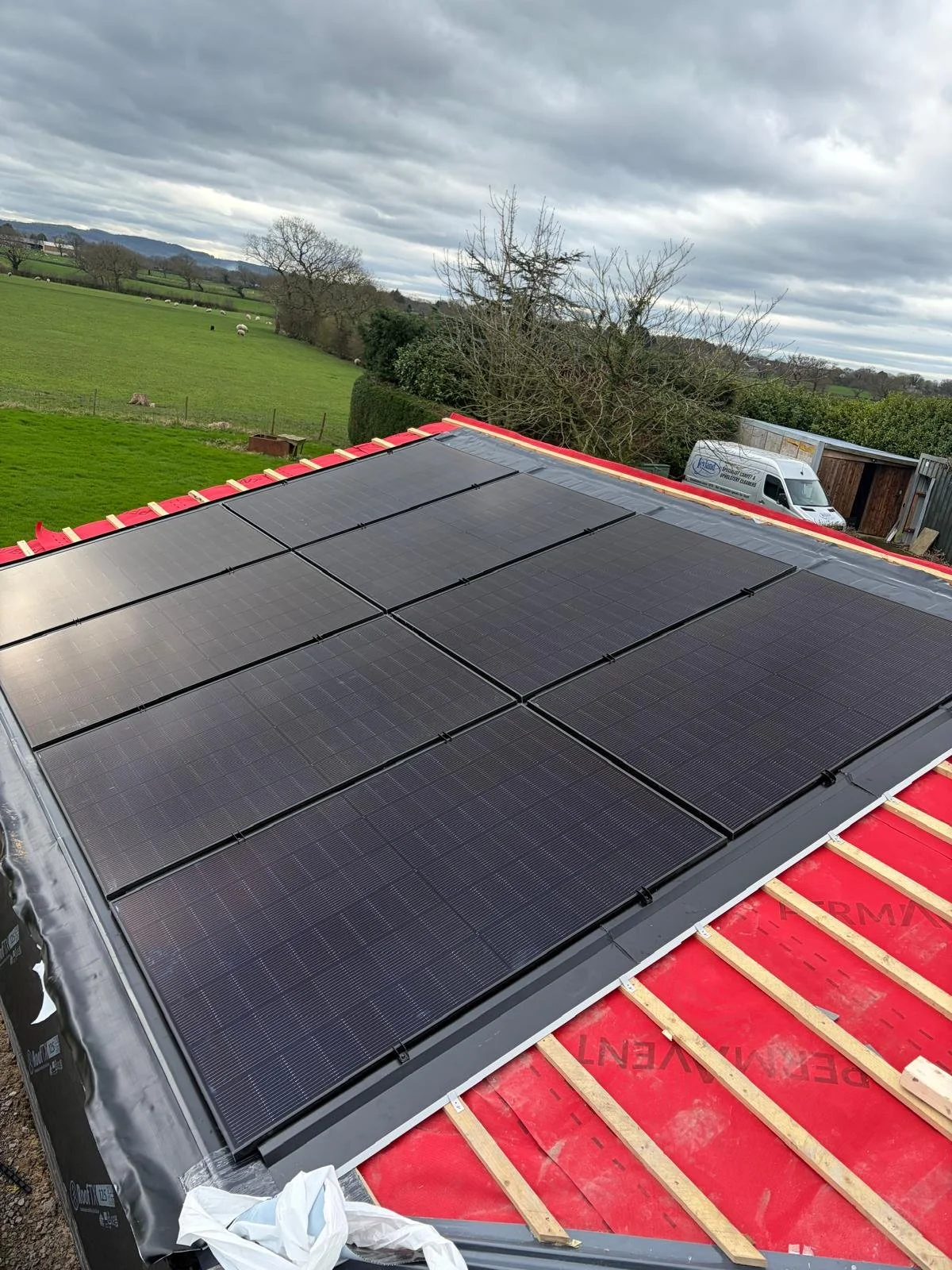 Roof with newly installed solar panels, black and rectangular, on red roofing under construction, with a scenic background of green fields, trees, and cloudy sky.