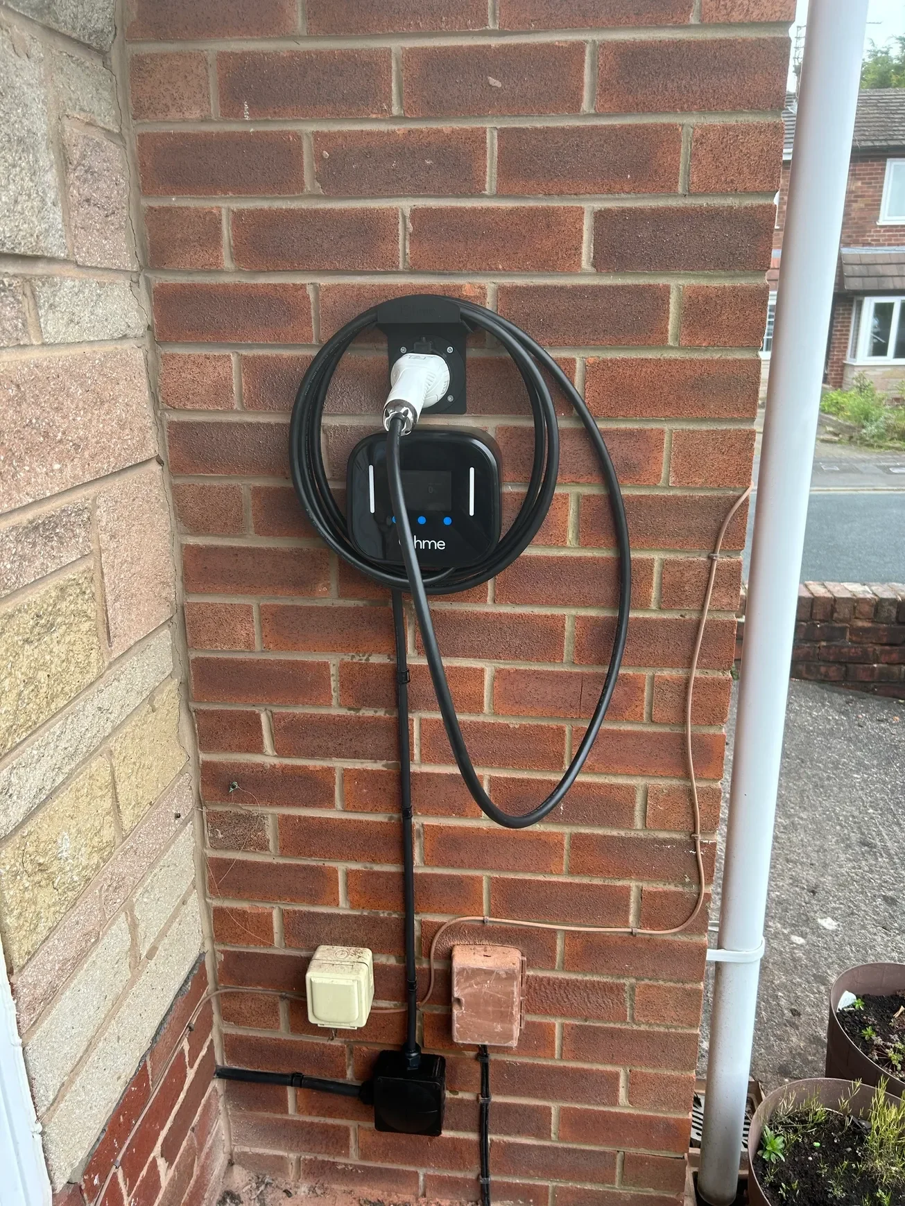 A brick wall with an electric vehicle charging station attached. The charging station is black with a white connector, multiple black cables, and some electrical outlets and wiring are visible below and around it.