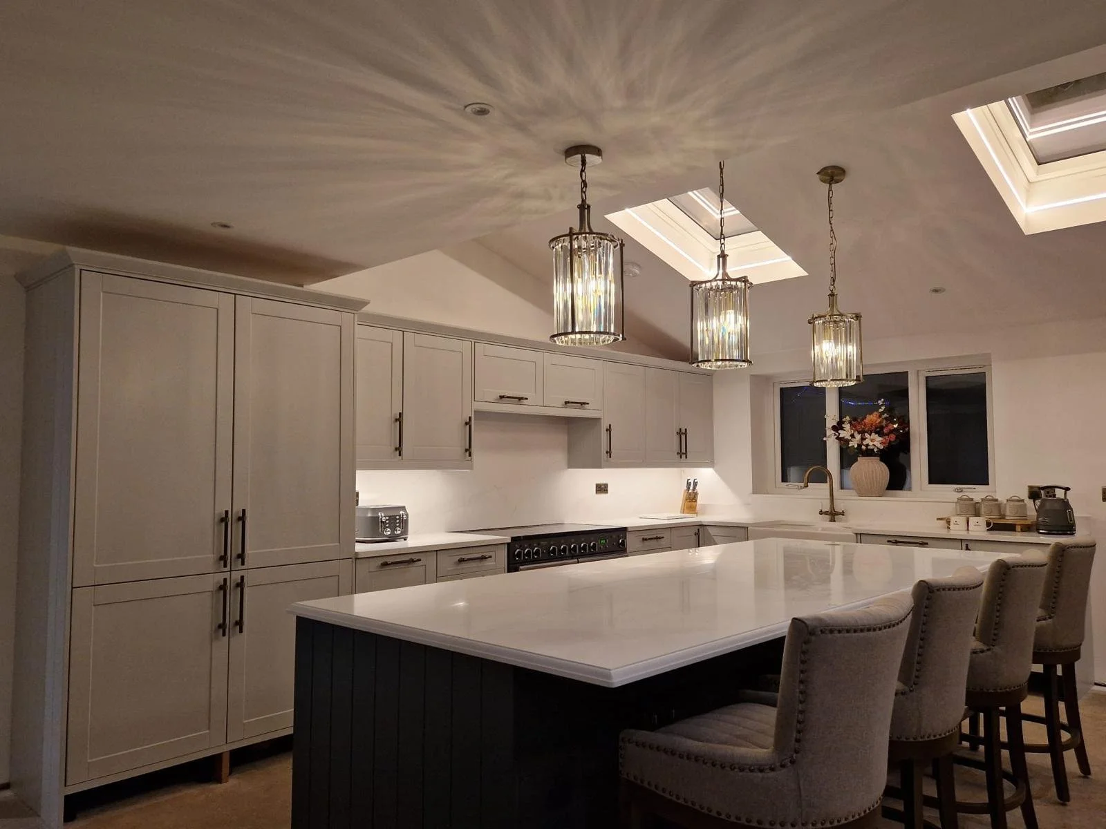 A modern kitchen with white cabinets, a kitchen island with a white countertop, three pendant lights hanging from the ceiling, a window with a vase of flowers, and bar stools around the island.