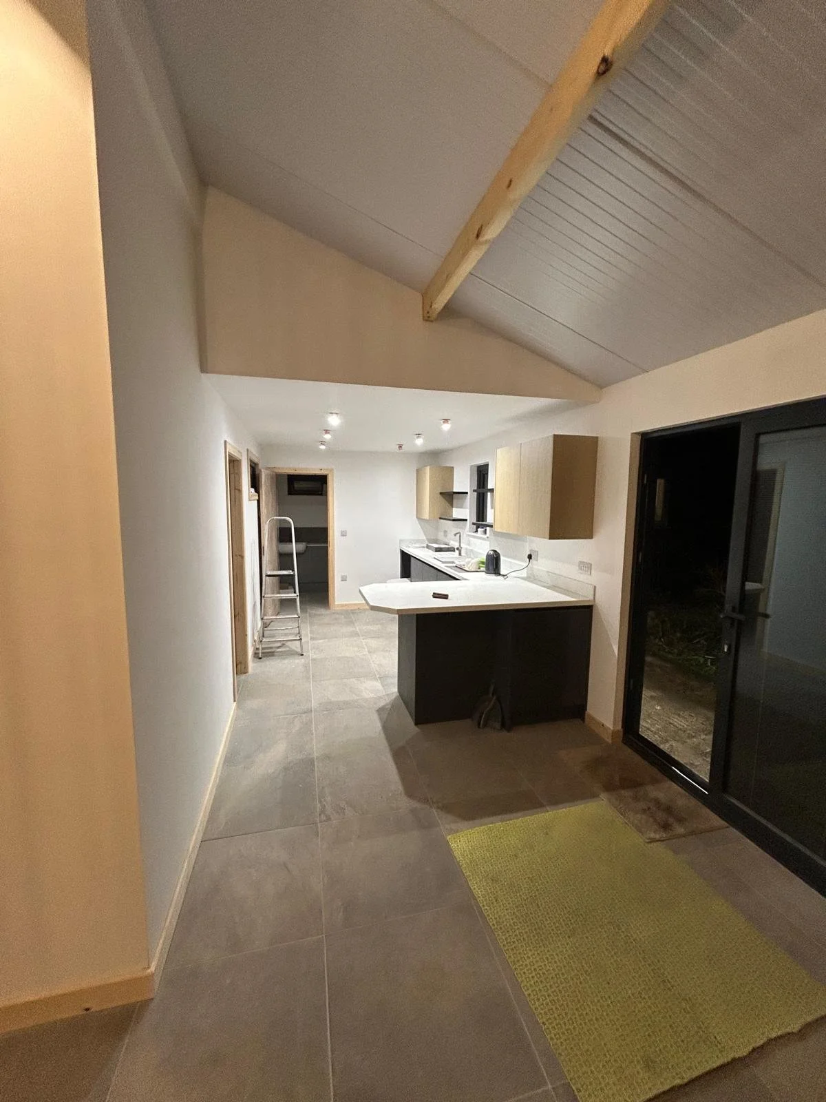 Modern kitchen with gray tile flooring, white countertops, wooden cabinets, black lower cabinets, and large black sliding glass doors leading outside. A step ladder is positioned at the far end near a small open space.