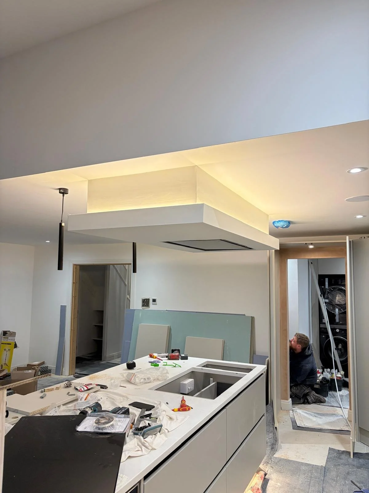 Interior of a house under construction or renovation, with workers installing cabinetry and lighting, and construction tools and materials scattered on the countertops.