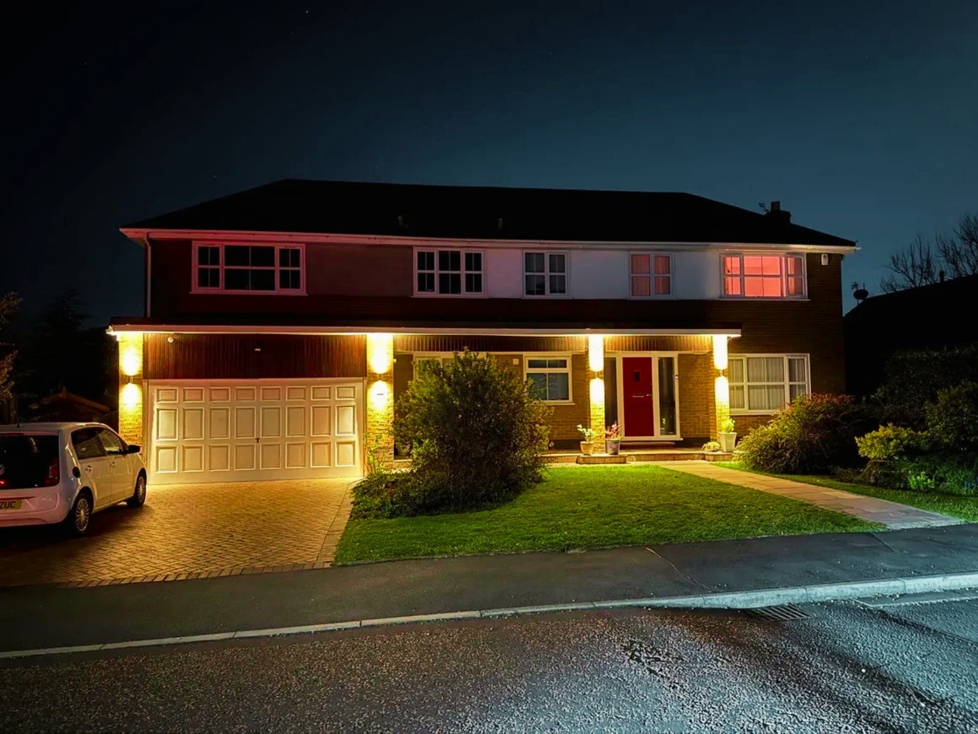 A two-story house illuminated at night with exterior lights. The house has a brick lower level with a white garage door and a red front door. The upper level has multiple windows with some showing pink or orange light. There is a tree and plants in the front yard, and a car is parked in the driveway. The street in front has wet pavement.