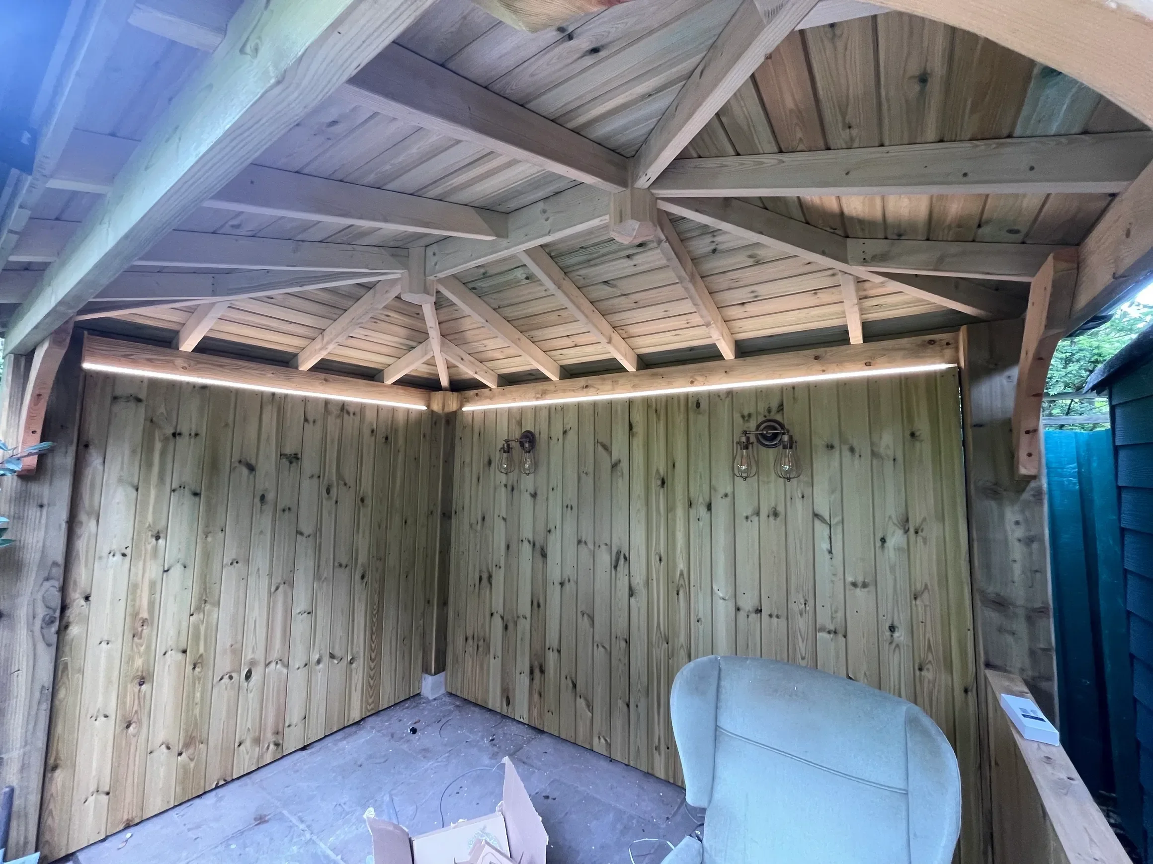 Interior of a wooden gazebo with a vaulted ceiling, wooden walls, and hanging light fixtures, under construction.
