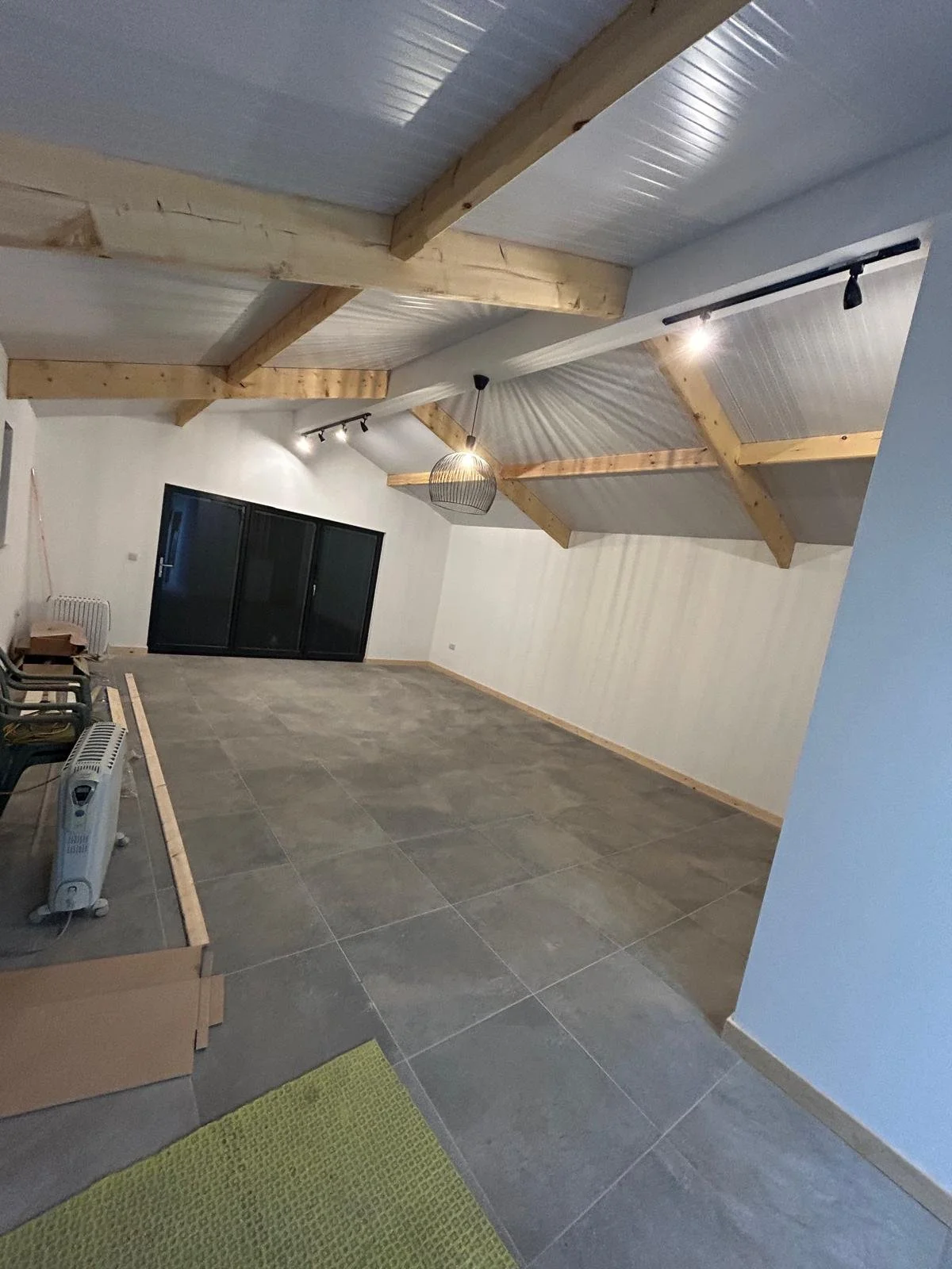 An unfinished room with a vaulted ceiling featuring wooden beams, white paneling, and installed track lighting. The floor is tiled, and there are chairs, a box, and a space heater in the corner. A large sliding glass door leads outside, and a green r