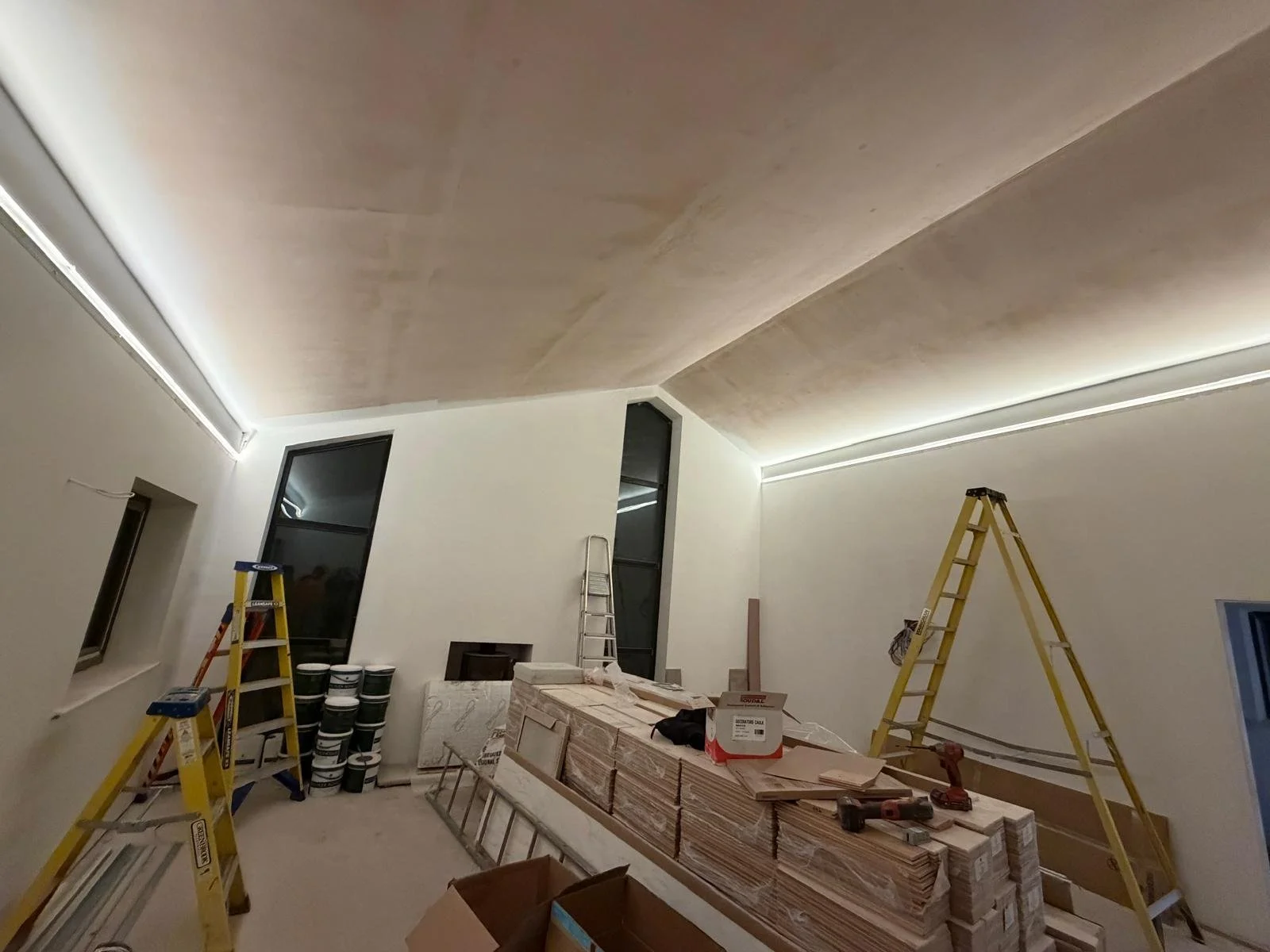 An unfinished room under renovation with ladders, construction materials, and tools, featuring a ceiling with hidden LED strip lighting.