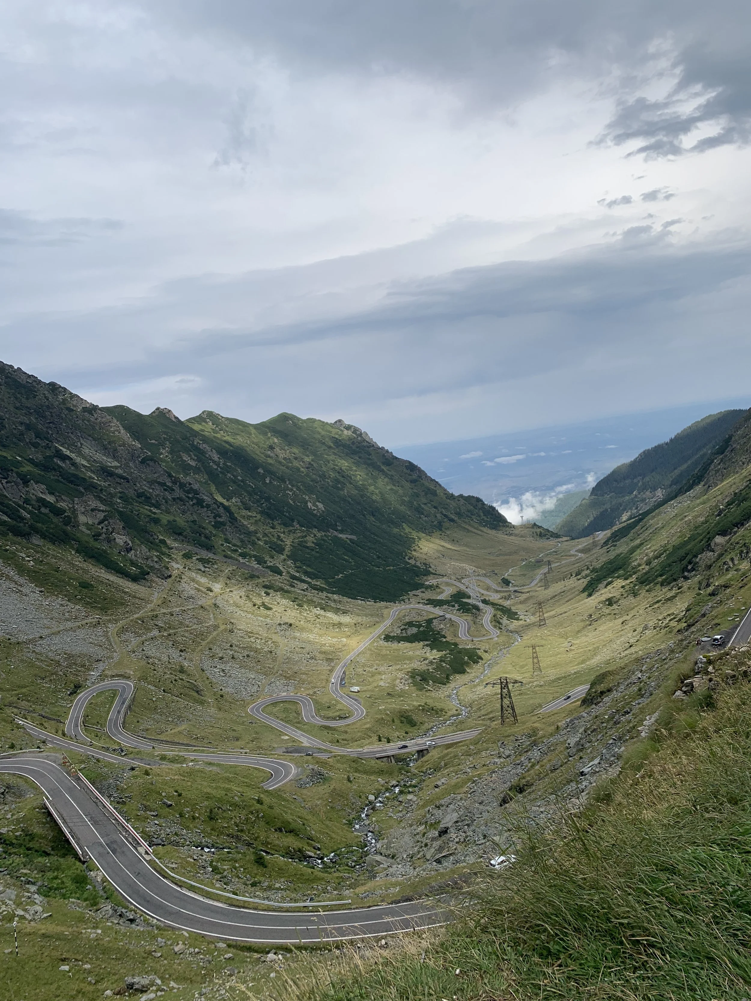A winding mountain road with sharp turns and switchbacks through green hills and rocky terrain, with steep slopes on either side and power lines crossing the valley, under a cloudy sky.