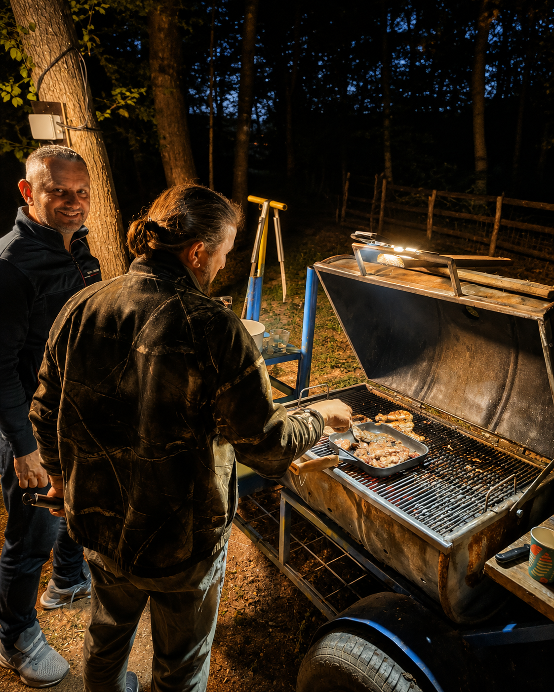 Two men are barbecuing chicken on a grill outdoors at night. One man is standing by the grill, turning chicken, while the other man stands nearby, smiling at the camera. The scene is illuminated by a small overhead light, with trees and a fence visible in the background.