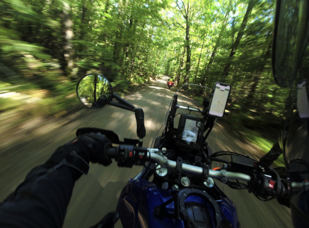 A motorcycle rider speeding through a forested trail with trees and greenery on both sides, viewed from the rider's perspective, with a rearview mirror and navigation device in view.