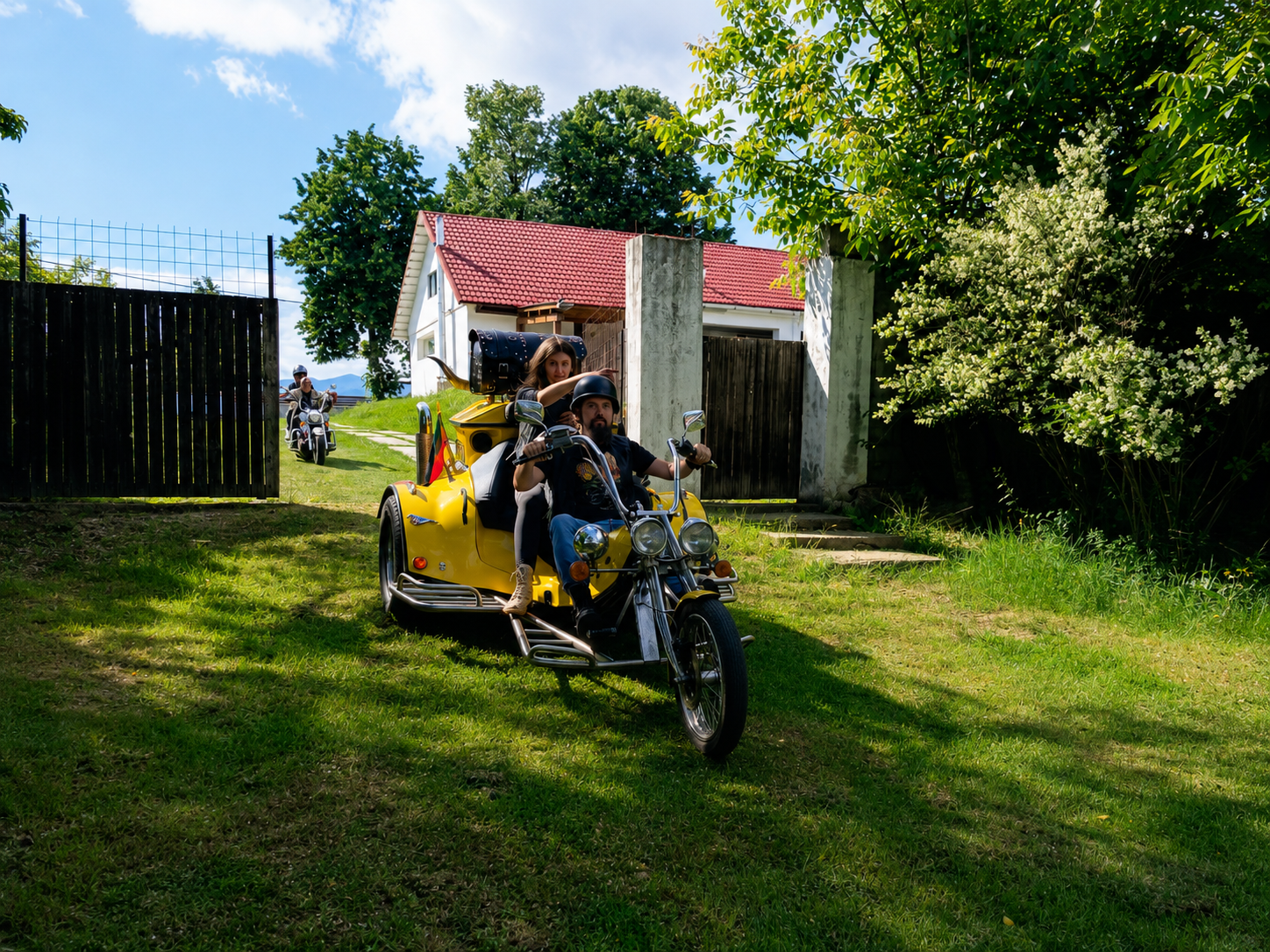 People riding a yellow motorcycle with a sidecar outside a house with a red roof and black gate, surrounded by trees and greenery on a sunny day.