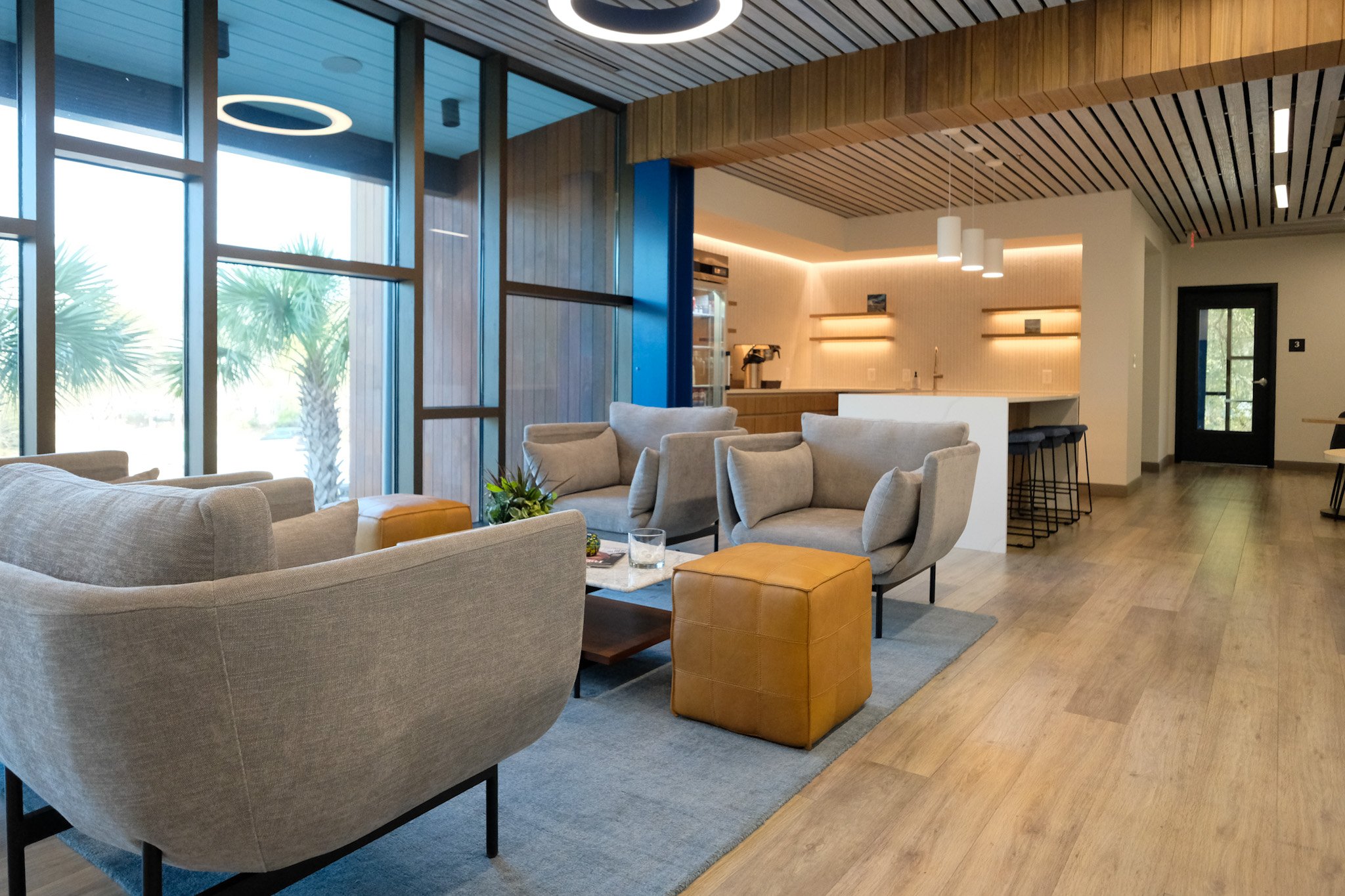 Modern lounge with beige and gray armchairs, a small coffee table, and yellow ottomans, large windows with palm trees outside, and a kitchen area in the background with bar stools.
