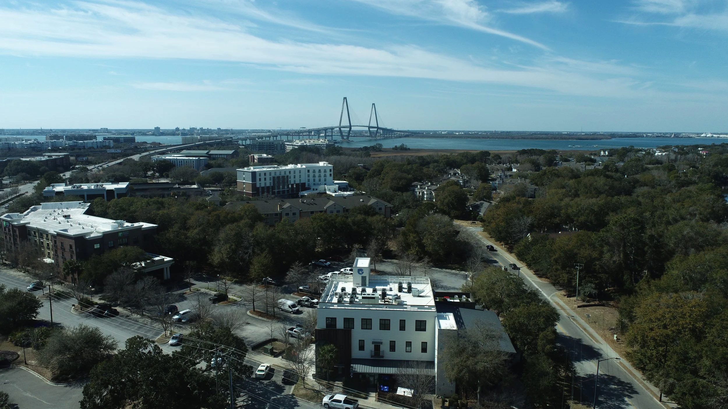 Aerial view of a coastal city with trees, roads, buildings, and a large bridge over a body of water in the background.