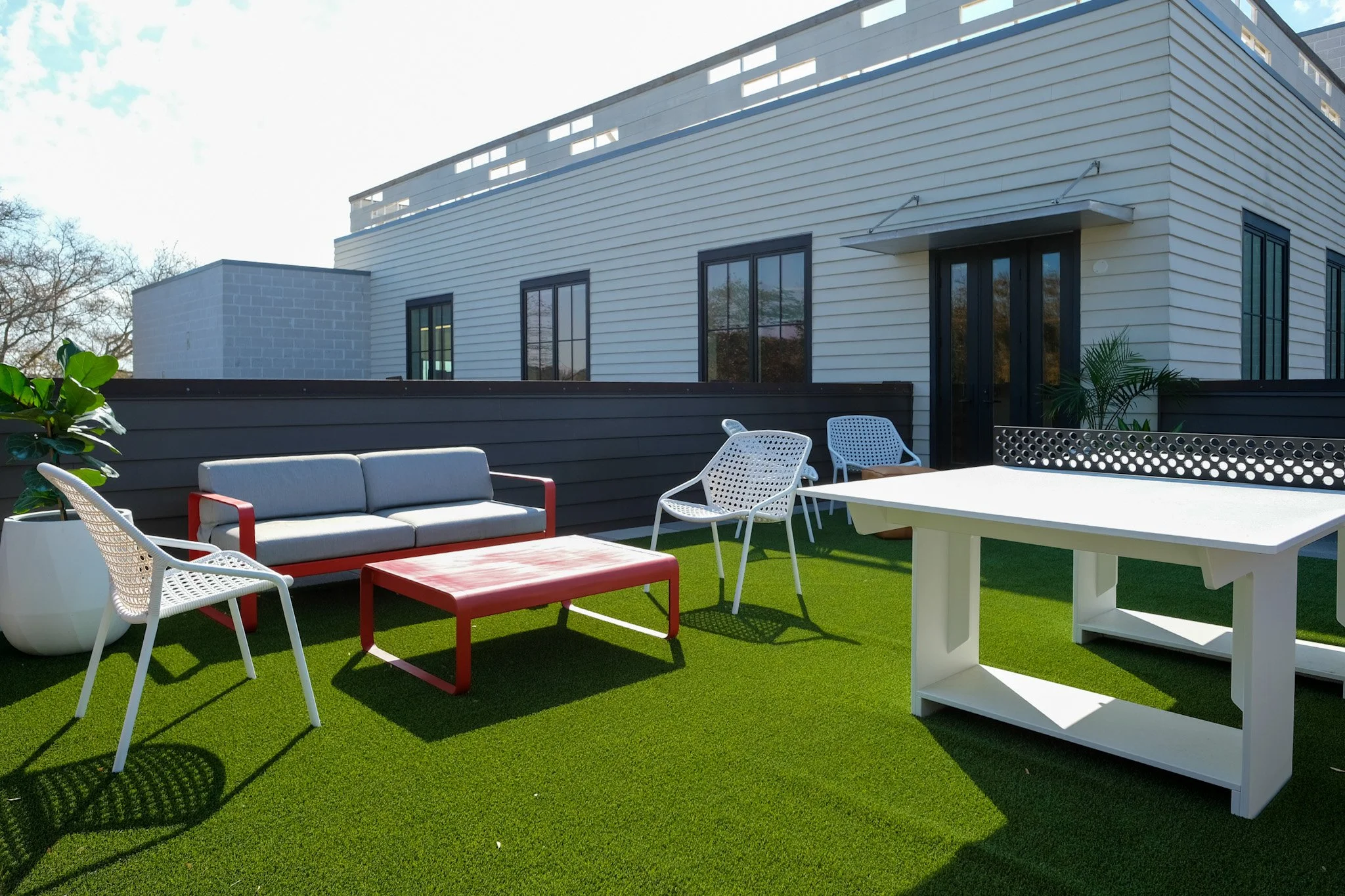 Modern rooftop patio with outdoor furniture, including a gray sofa with red accents, white chairs, a red coffee table, and a white table on artificial grass, surrounded by a building with large windows.